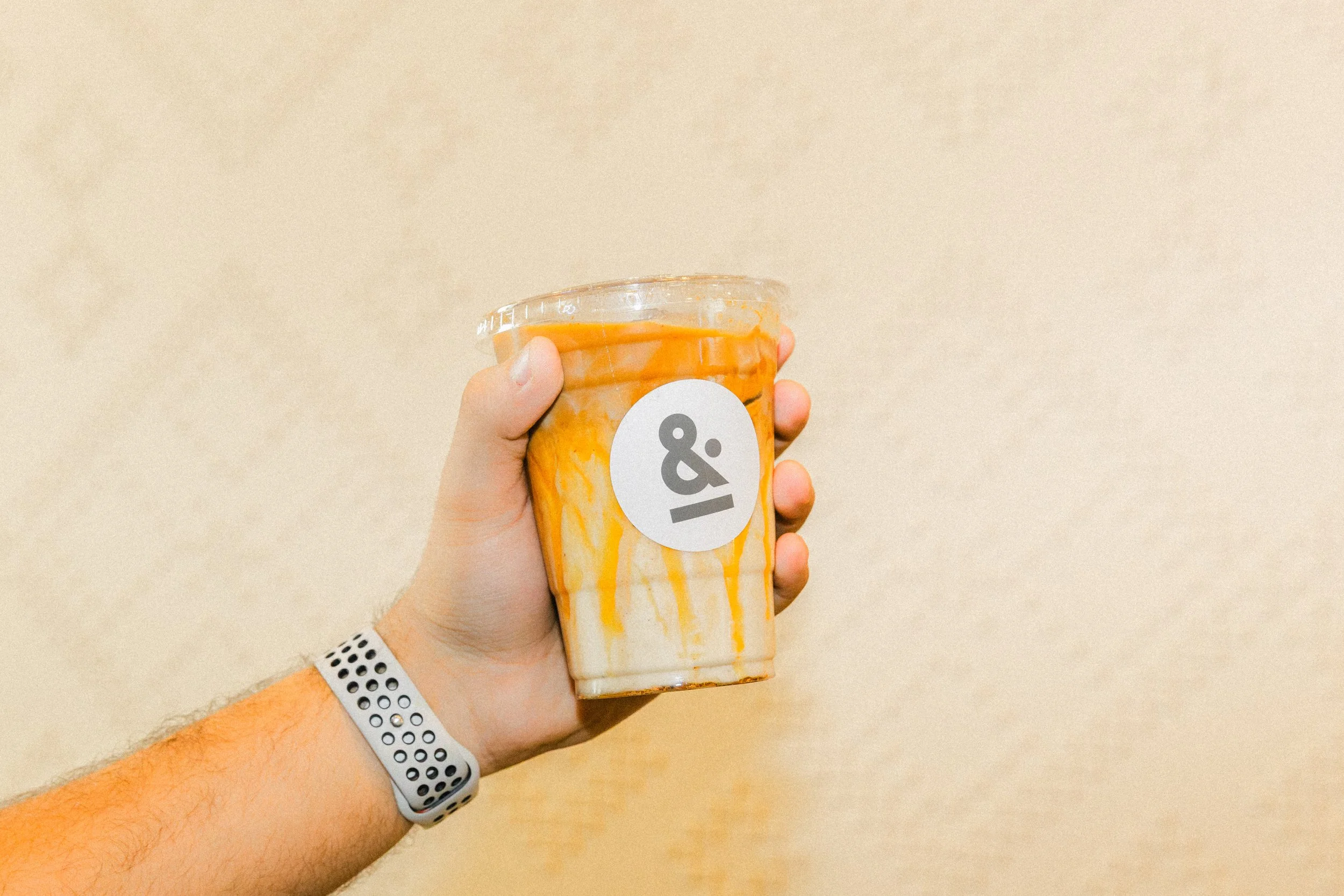 A person's hand holding a plastic cup of iced coffee or milk tea with orange and white swirls, and a white logo with black text '&' on the front. The person is wearing a white wristband with black polka dots.