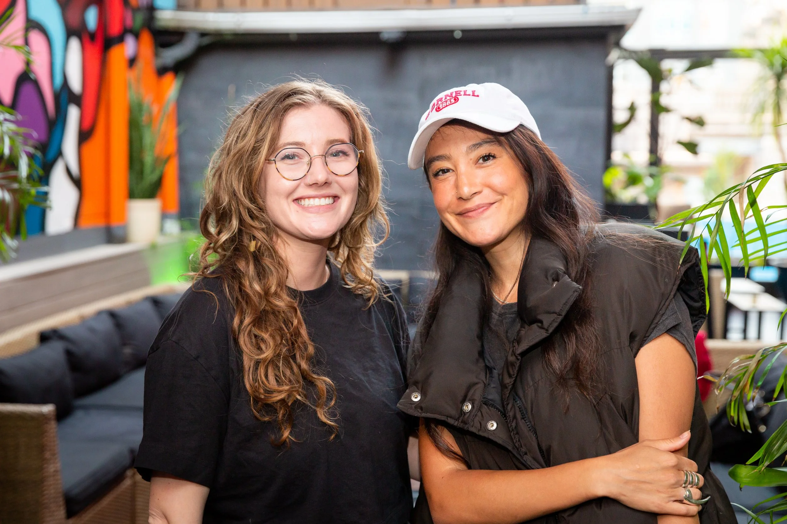 Two women smiling, one with curly red hair, glasses, and a black t-shirt, and the other with long dark hair, a white cap, and a black puffy vest, standing close together in a colorful indoor setting with plants and decorative wall art.