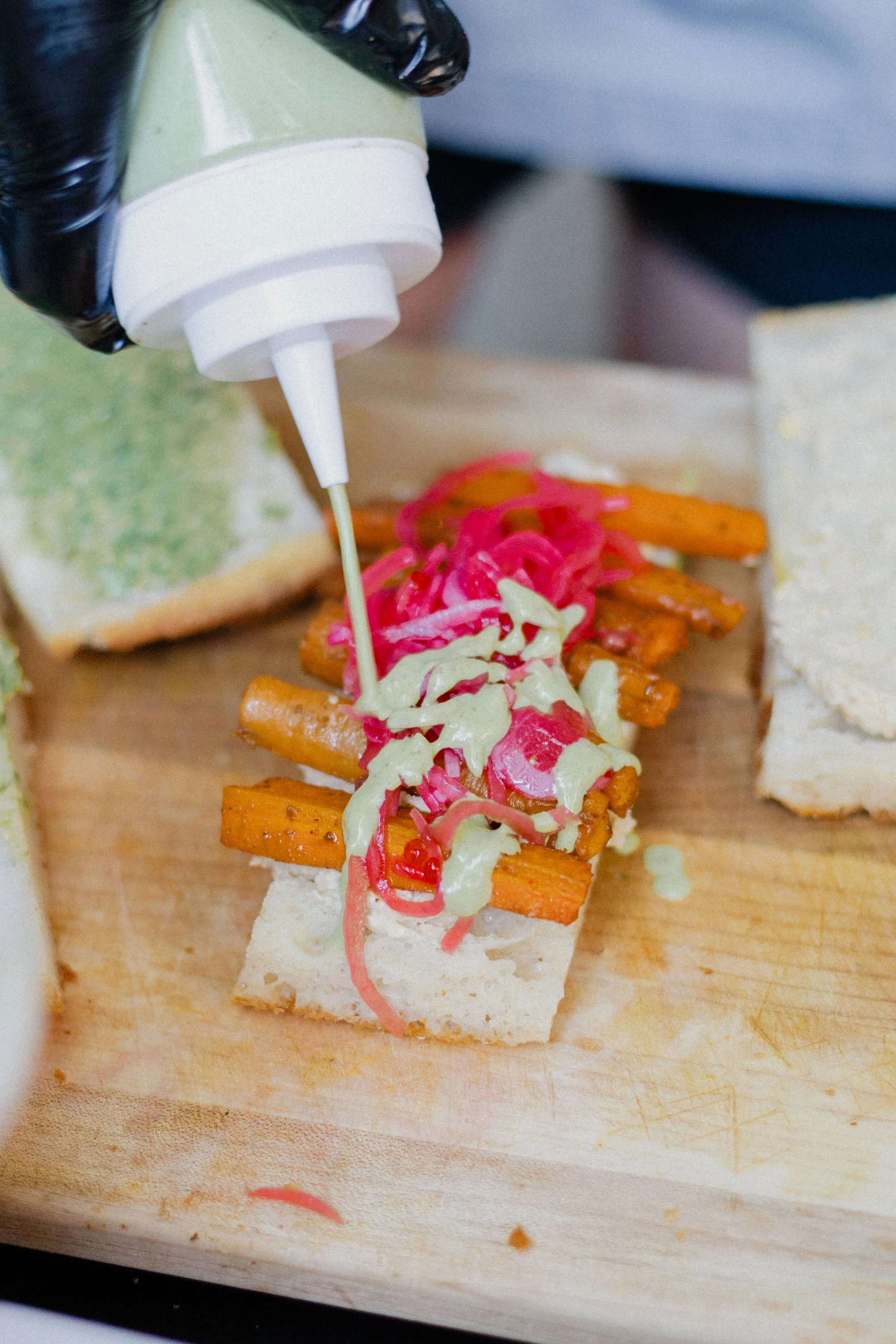 Sauce being drizzled on sweet potato fries topped with pickled vegetables on a piece of bread, on a cutting board.
