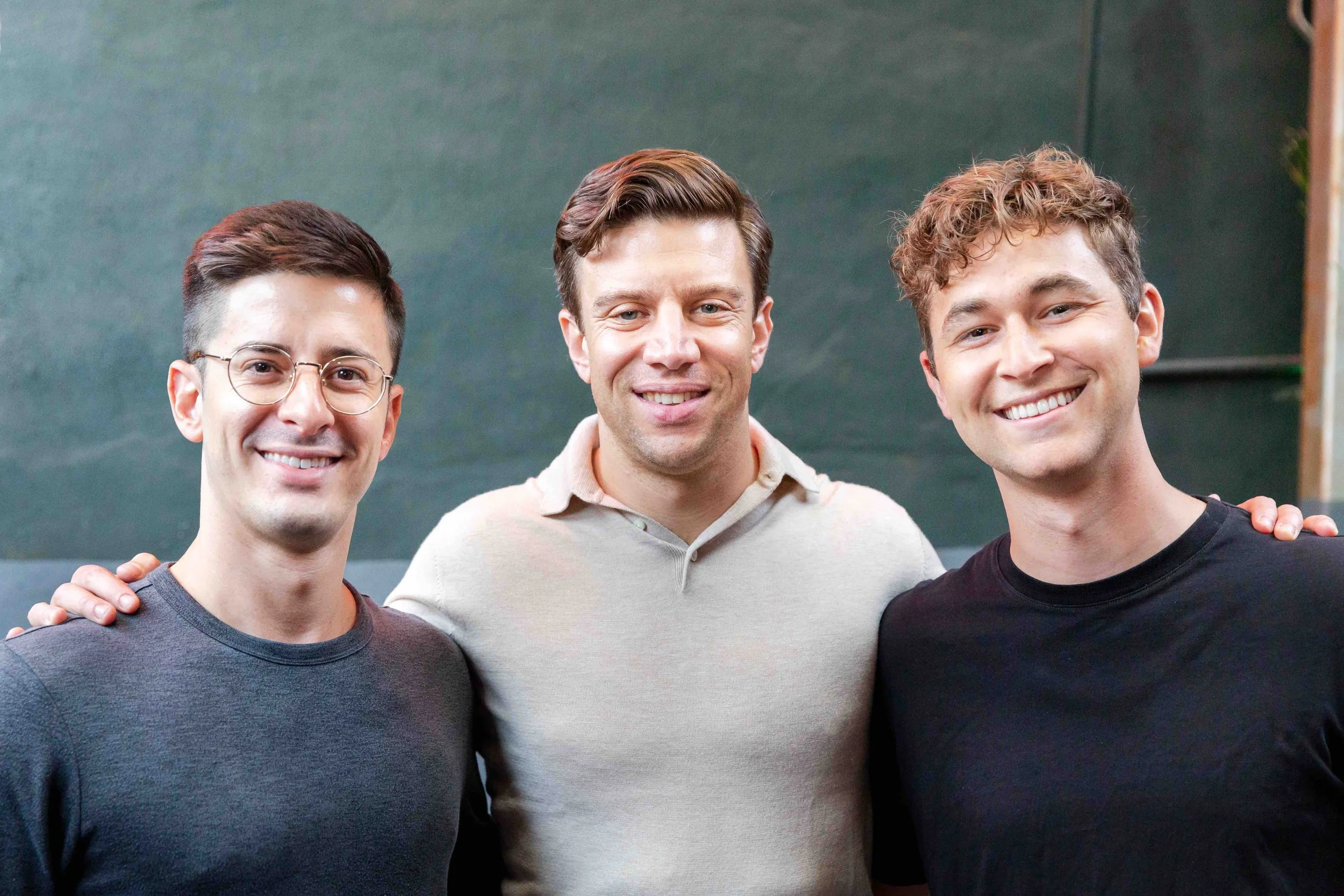 Three young men smiling and standing close together in front of a dark green background, with one man in the middle having his hands on the shoulders of the other two.