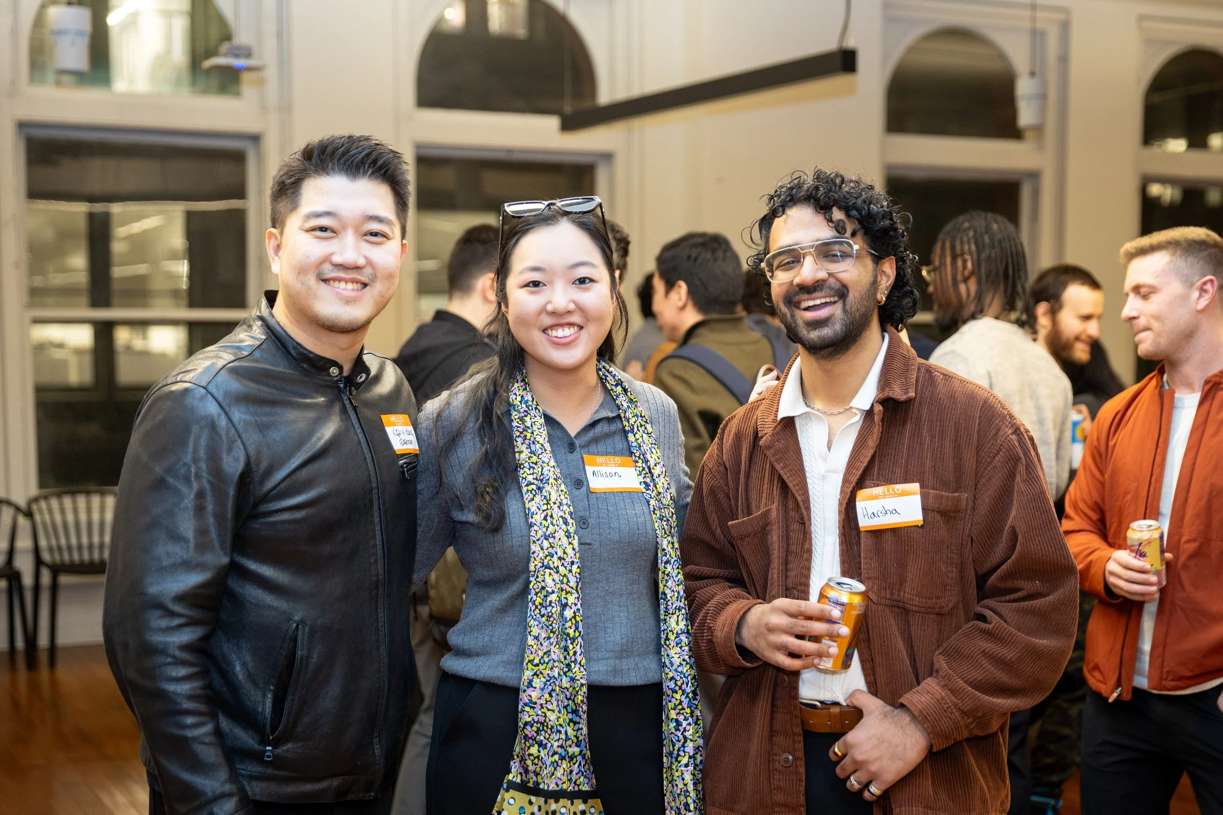Three people smiling at a social gathering, wearing name tags, with additional people in the background in an indoor setting.