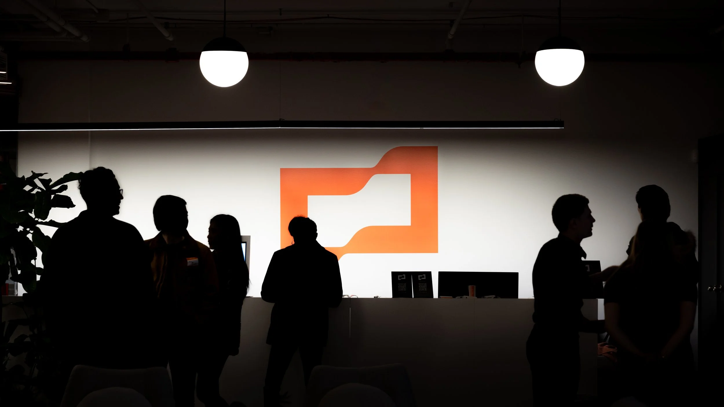Silhouettes of people standing and talking in front of a backlit wall with a logo, in a dimly lit room with hanging lights.