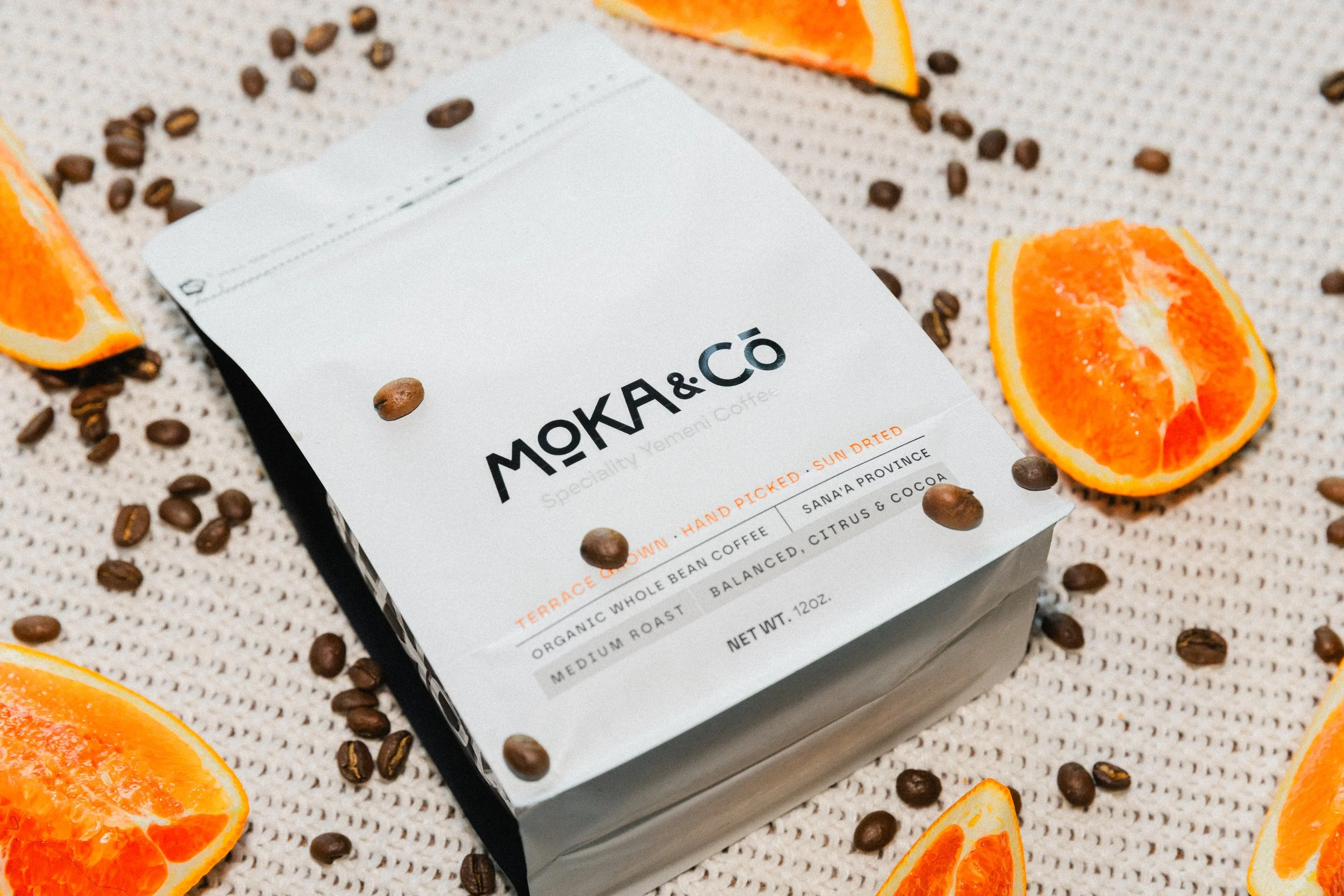 A white coffee bag labeled 'MOKA & Co Specialty Yemeni Coffee' surrounded by orange slices, coffee beans, and scattered coffee beans on a textured surface.