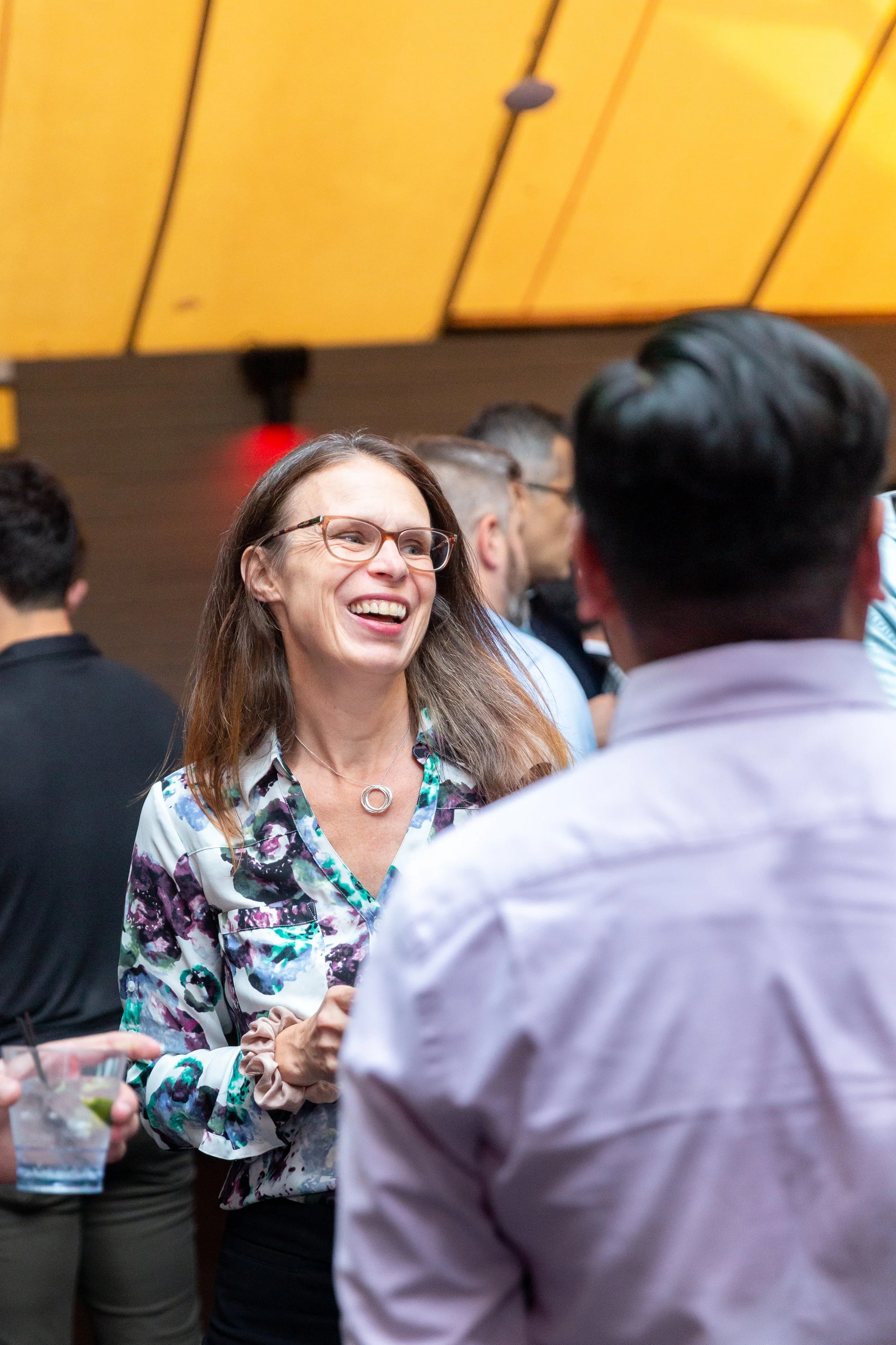 A woman with glasses and a floral blouse smiling and engaging in conversation at a social event.