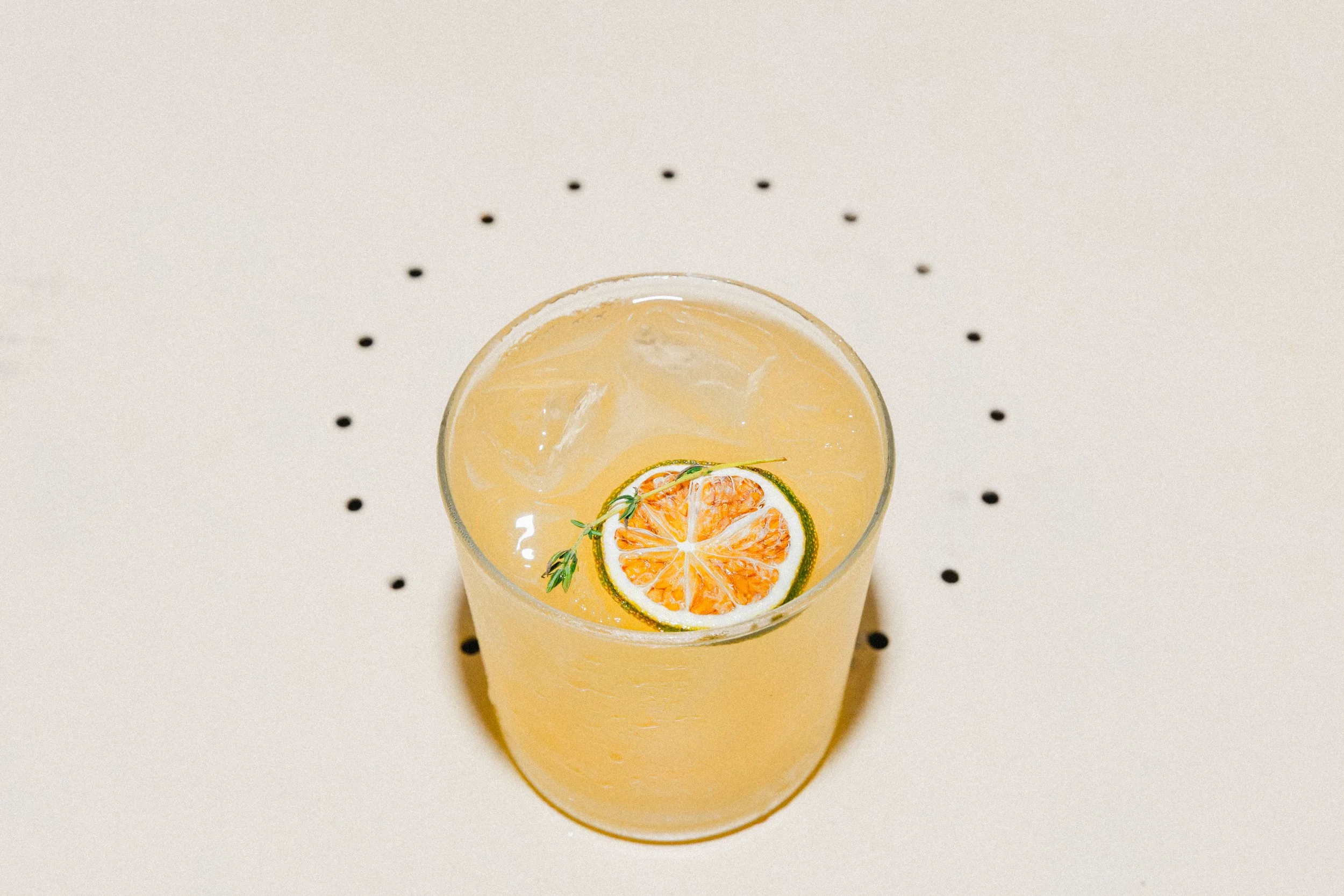 A glass of yellow citrus drink with ice, garnished with a slice of lime and a sprig of thyme on top, placed on a white surface with black dotted pattern around it.