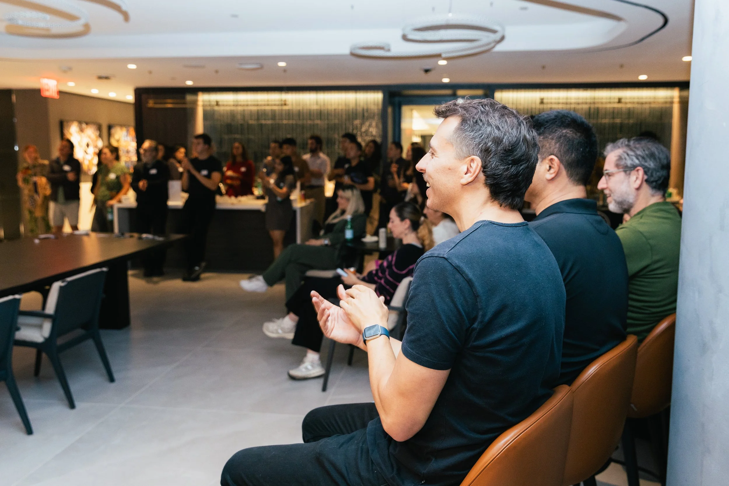 People attending a presentation or event in a modern indoor venue, with some seated and others standing near a bar area, engaging and smiling.