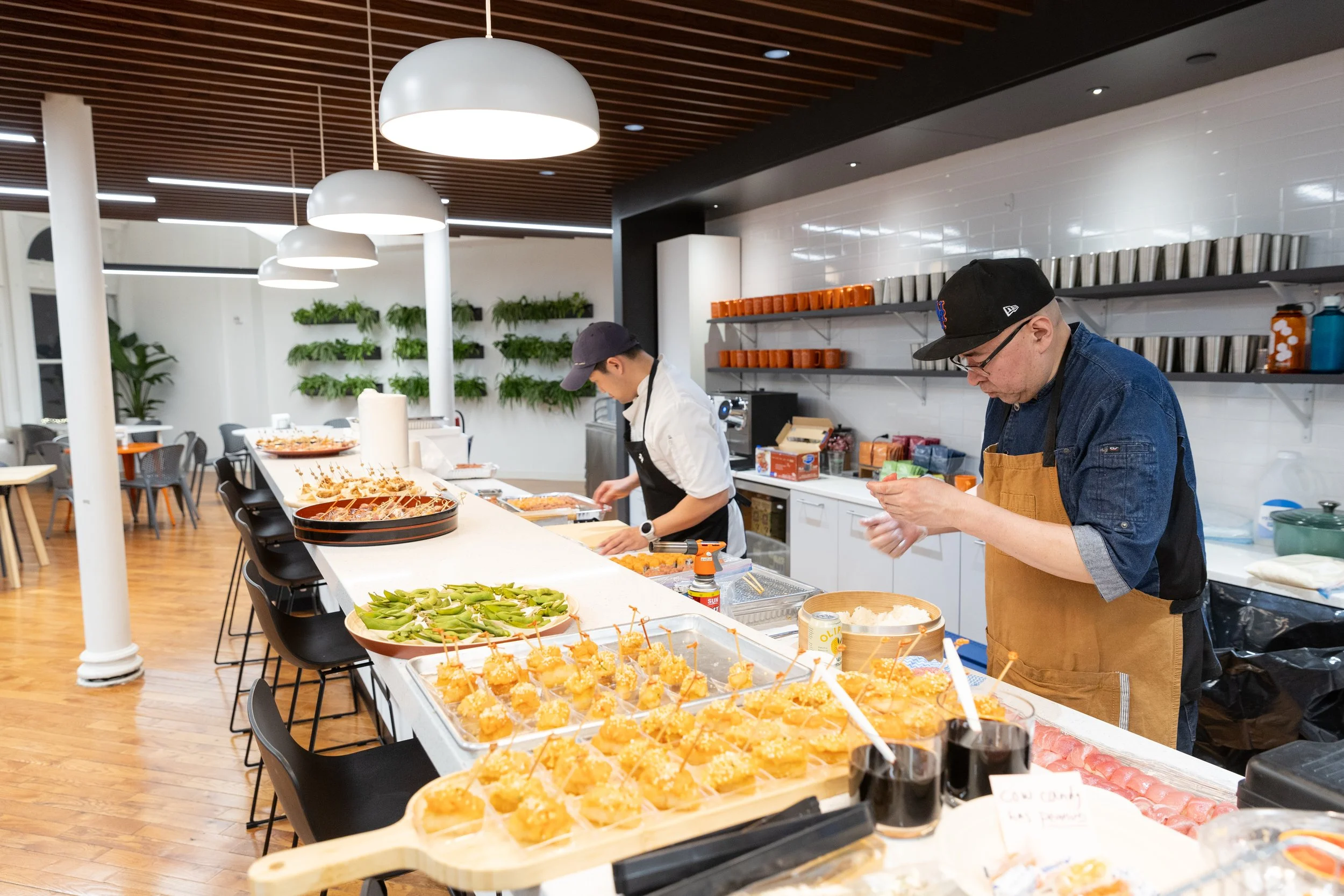 Two chefs preparing food at a modern restaurant counter with various appetizers like skewers, vegetables, and dips, in a well-lit space with wooden floors and decorative plants.