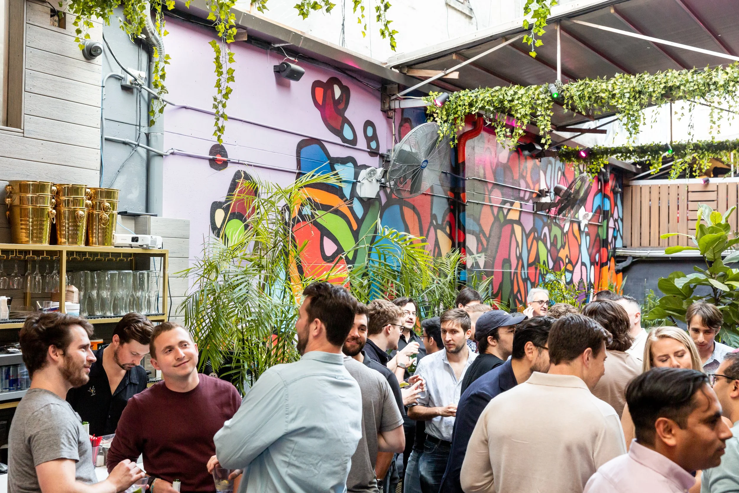 Crowd of people socializing at an indoor party with graffiti wall art, green plants, and hanging greenery.