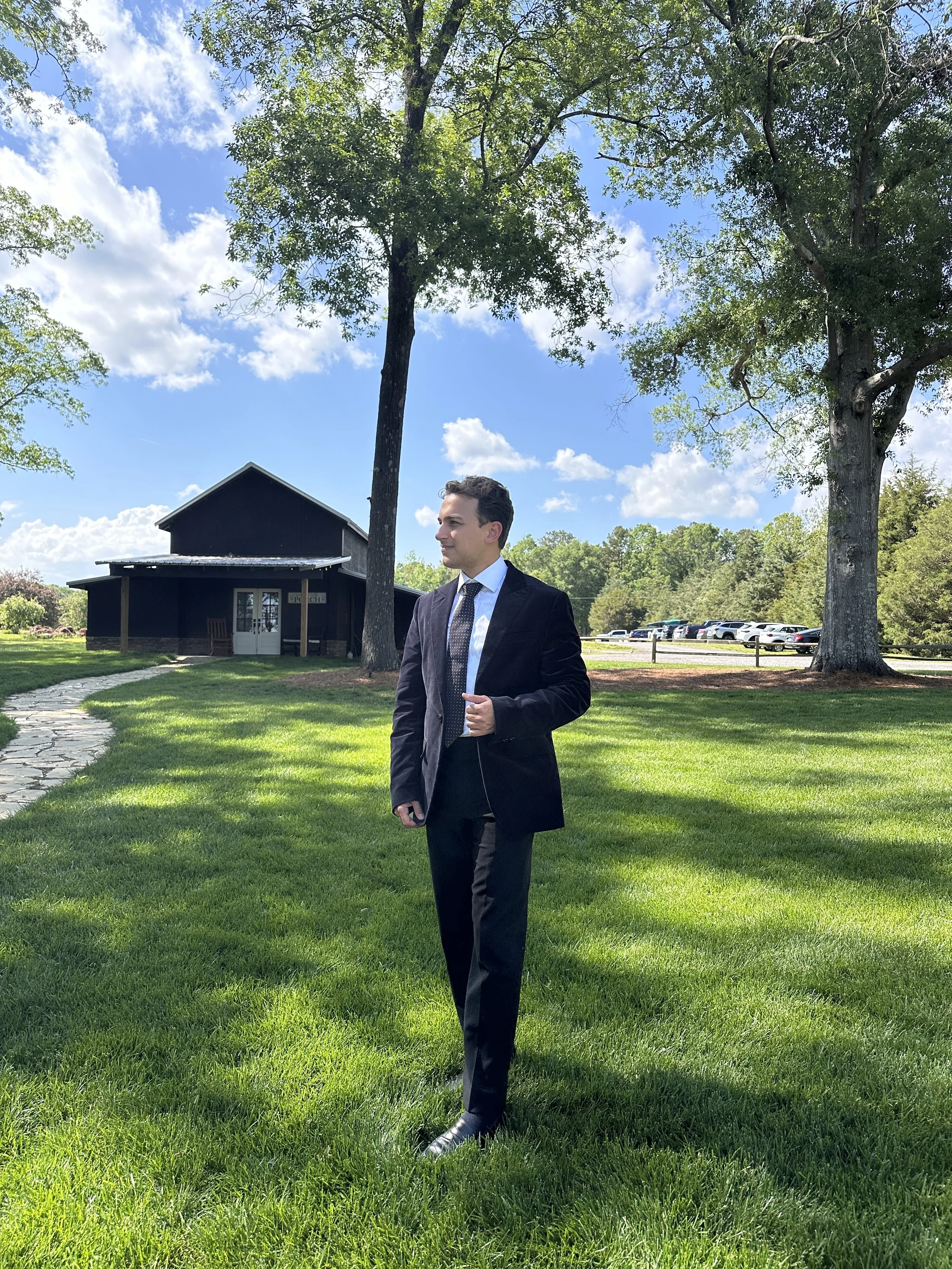 A man in a black suit and tie standing on a grassy lawn under large trees with a black building and parked cars in the background on a sunny day.