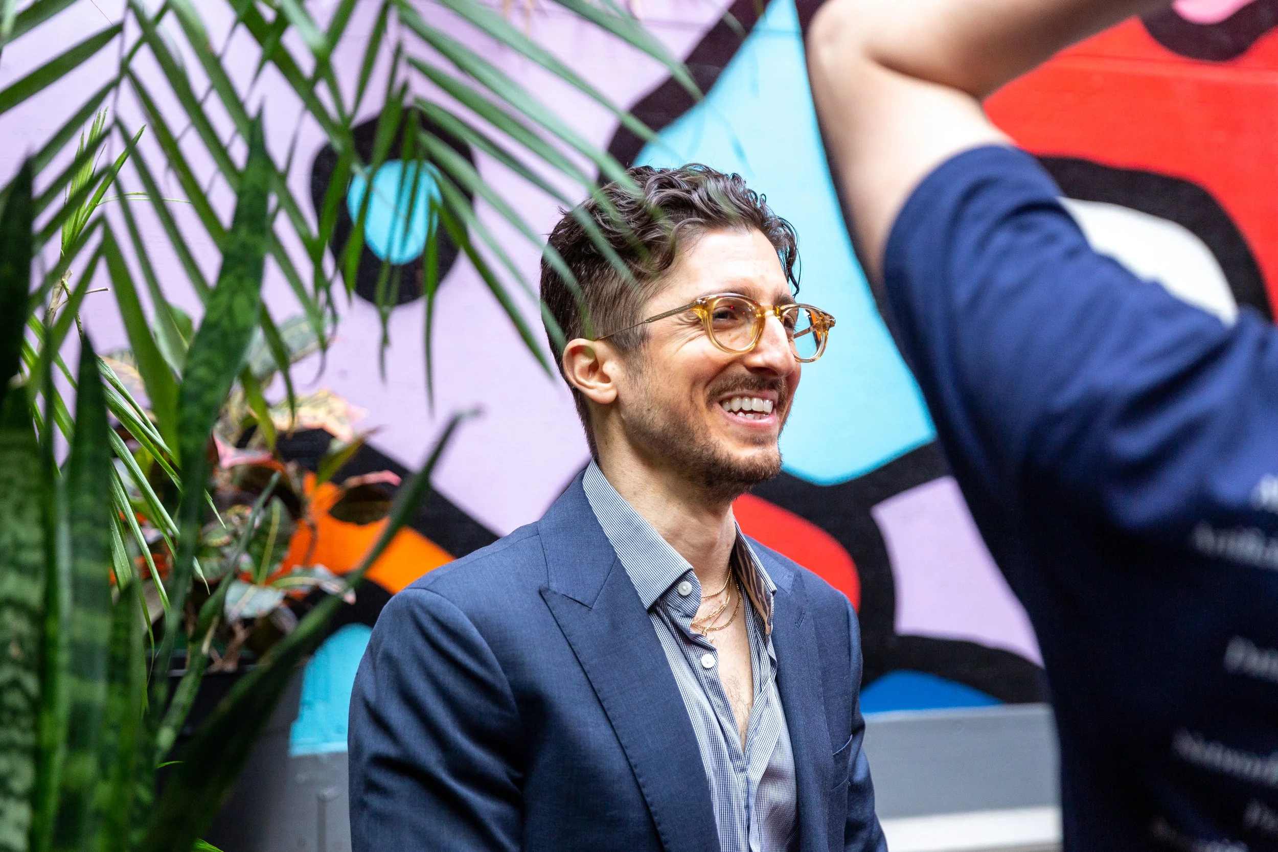 Man with curly hair, glasses, and a beard wearing a blue suit smiling in front of a colorful, geometric graffiti wall, with a person in a red shirt reaching over in the foreground.