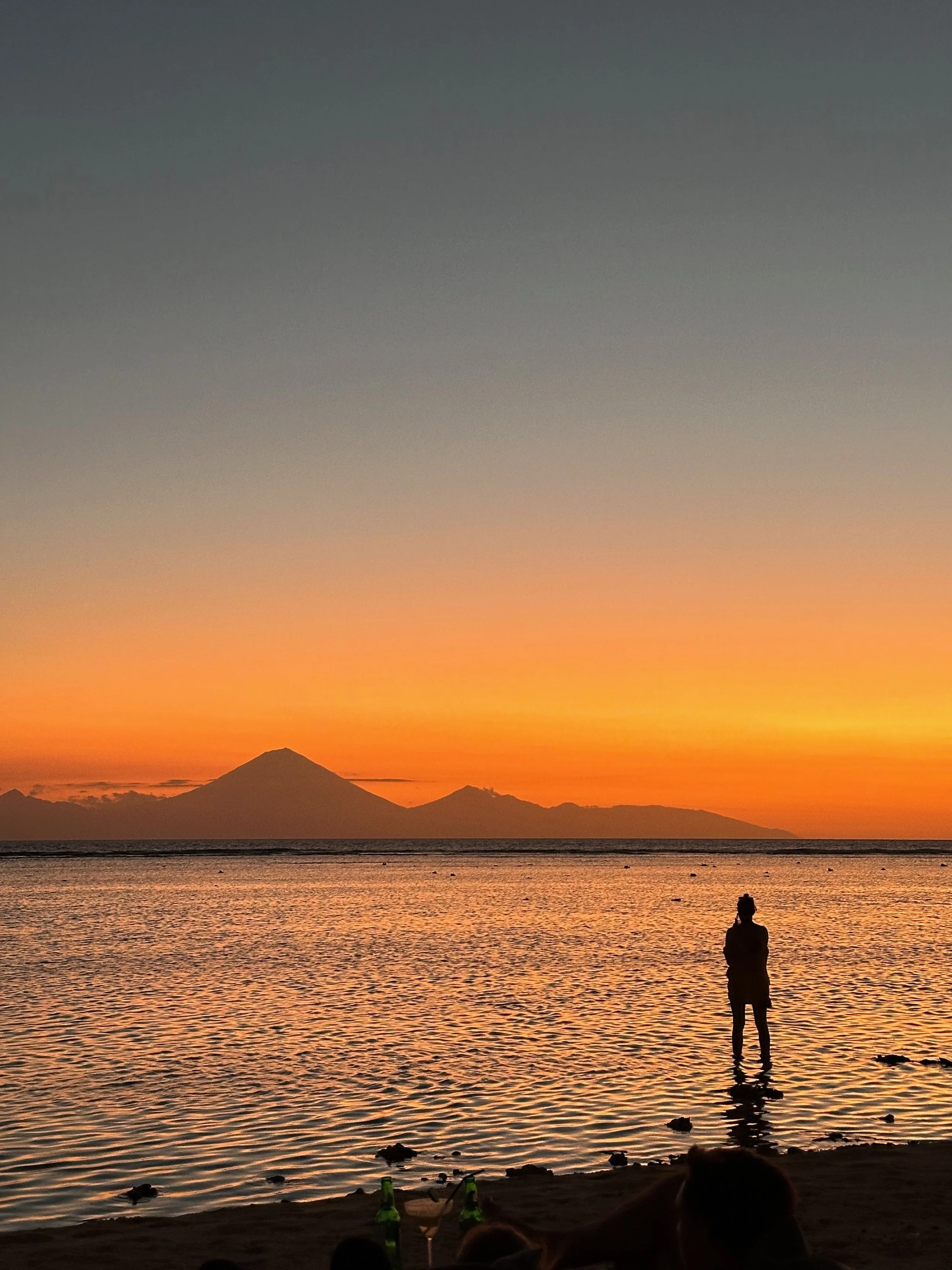 A person standing in shallow water at sunset, with Mount Fuji in the background and a colorful sky overhead.