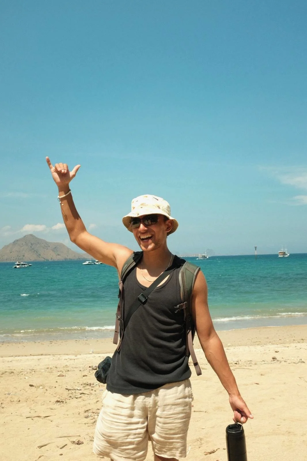 A man on a beach, smiling, wearing sunglasses, a beige hat, a black sleeveless shirt, beige shorts, and a backpack, holding a black water bottle, with a blue sky, ocean, and boats in the background.
