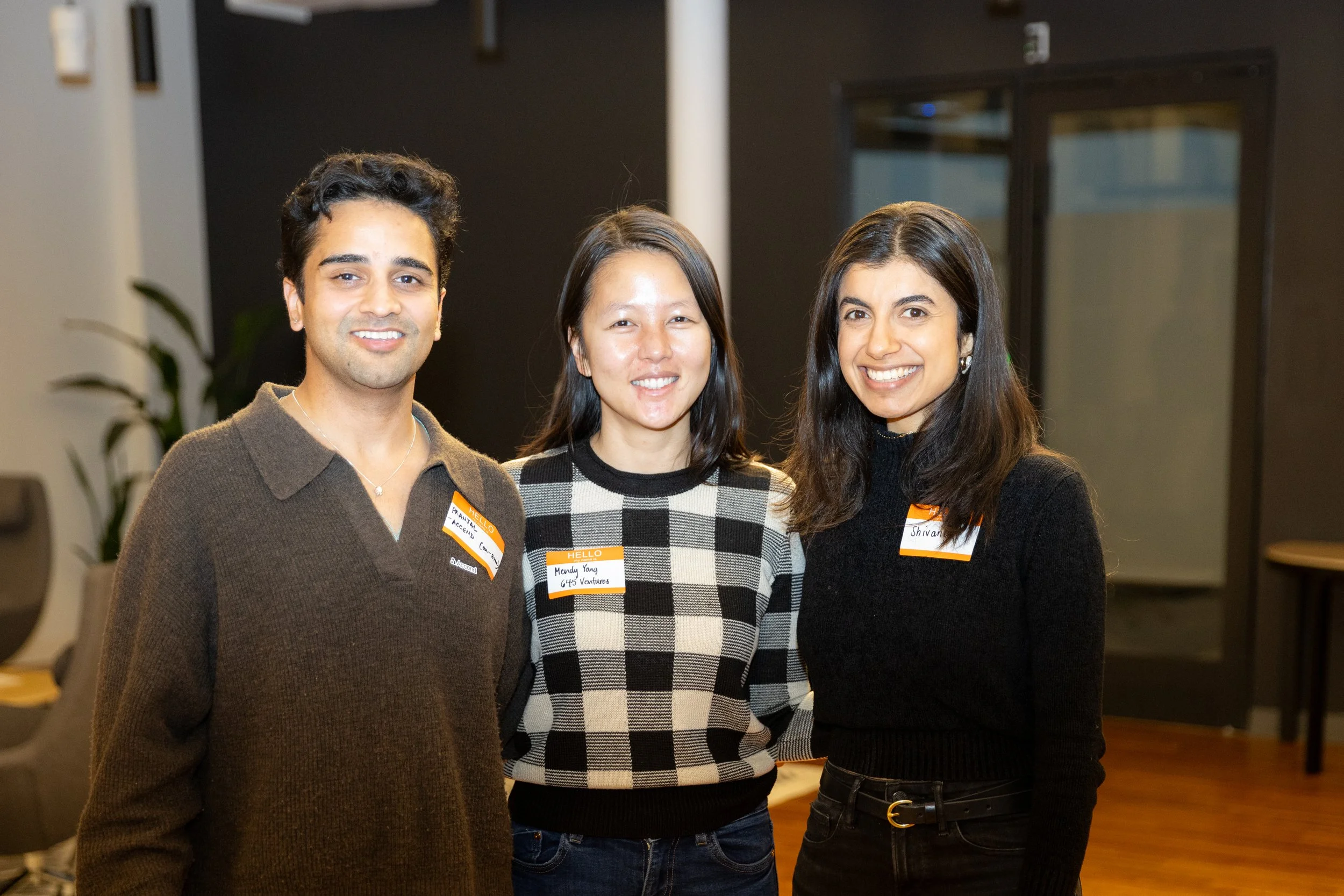 Three smiling people standing indoors, wearing name tags on their clothes.