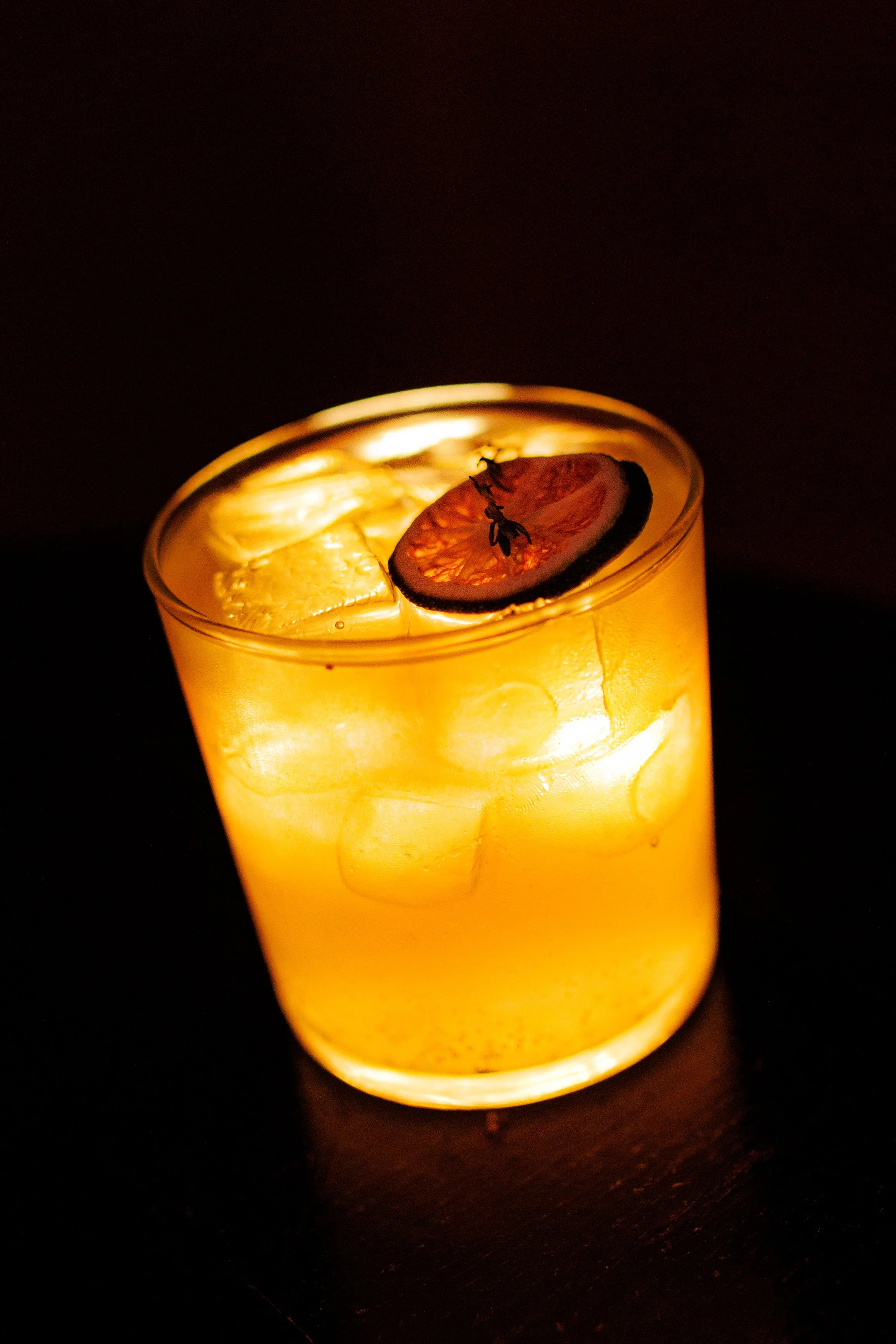 A glowing yellow cocktail in a clear glass with ice, garnished with a slice of blood orange and a small sprig of thyme, against a dark background.