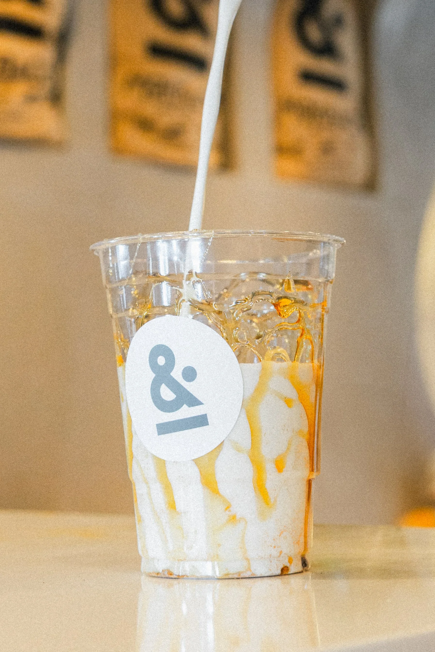 A clear plastic cup filled with ice and a creamy beverage, with caramel drizzle on top, being topped off with whipped cream from a white straw, on a light-colored surface, with blurred background signs.