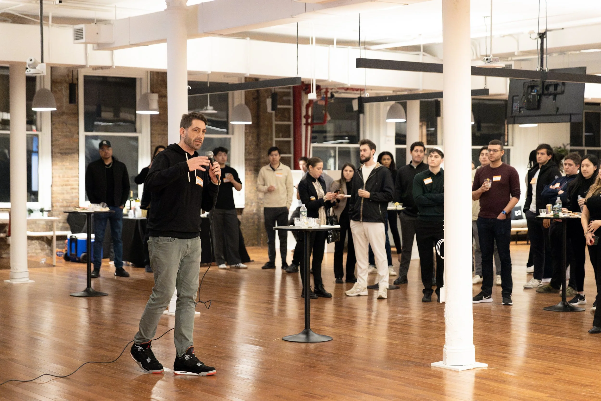 Man speaking into a microphone at an indoor networking or social event with attendees listening in a modern space with wooden floors, large windows, and exposed brick walls.