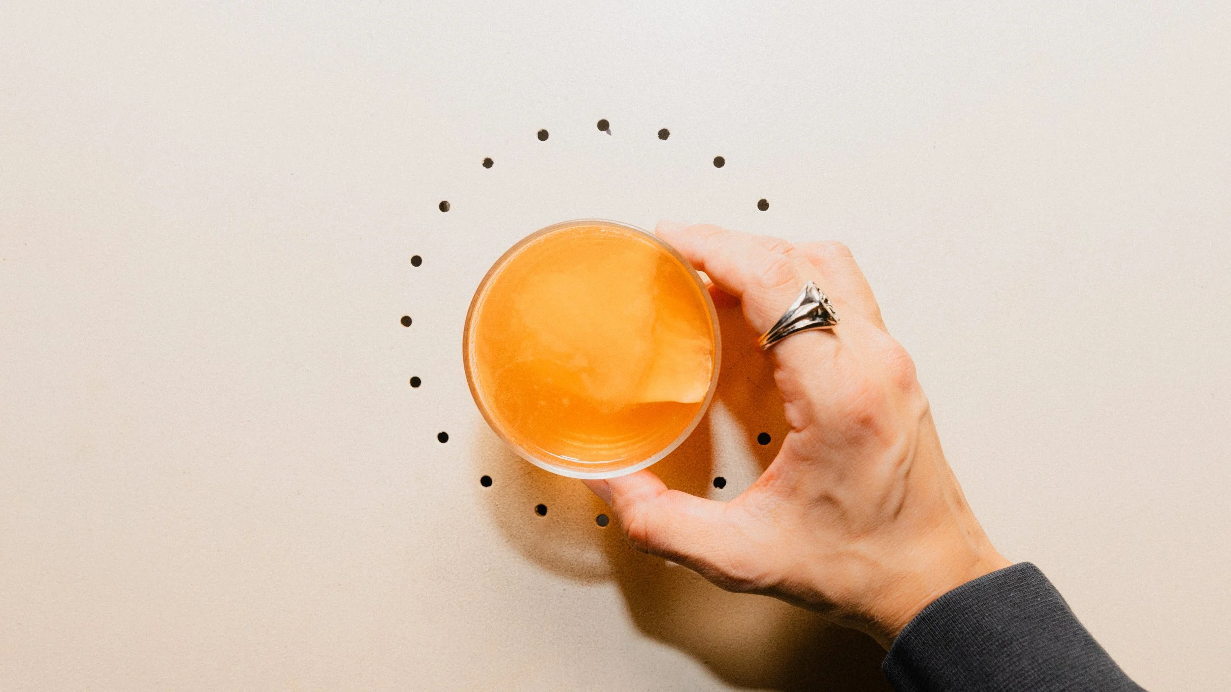 A hand holding a glass of orange-colored beverage over a white surface with black dotted circle design.