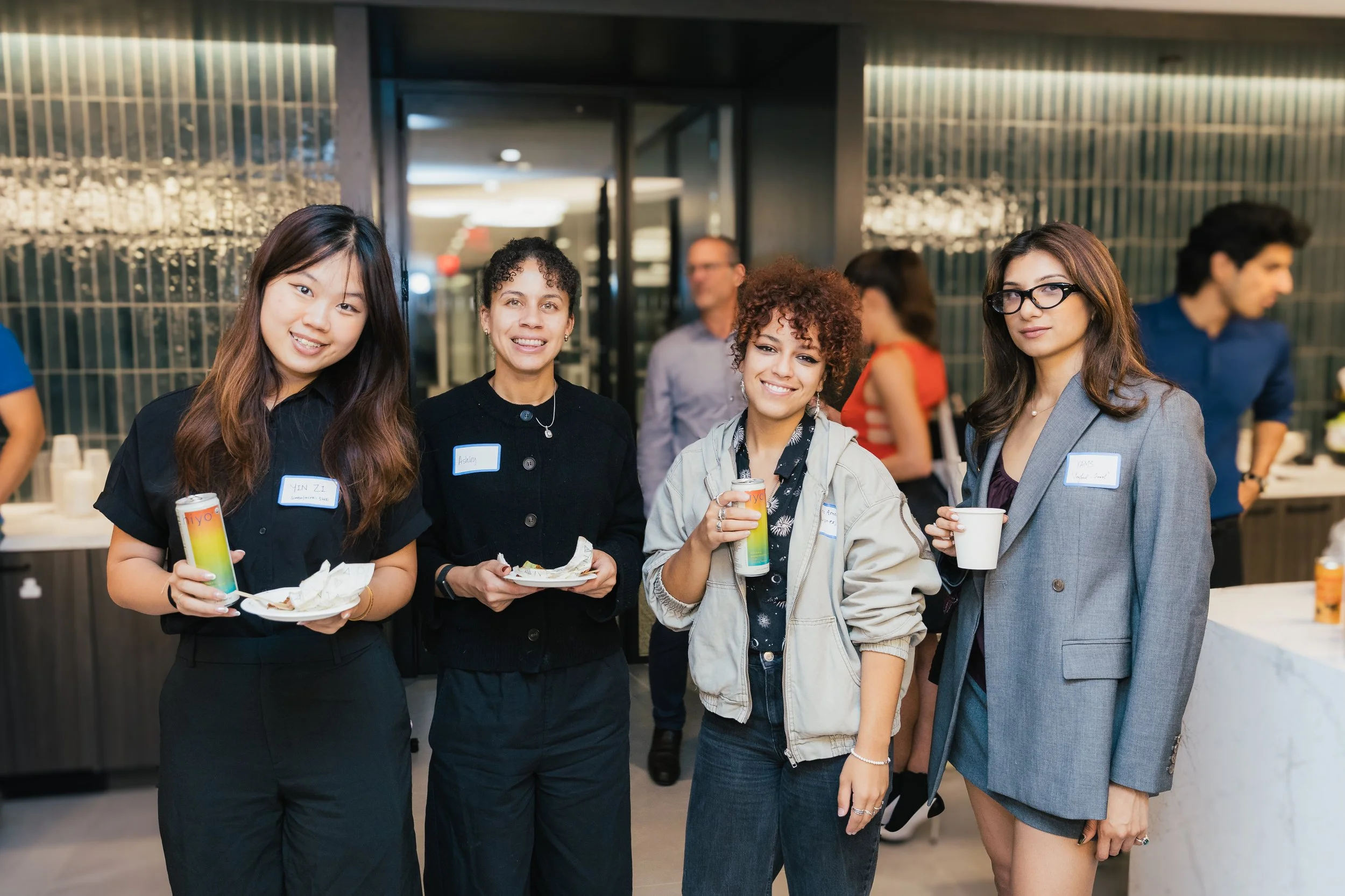 Four women standing together at an event, holding drinks and plates of food, smiling at the camera. They are in a modern indoor setting with other people in the background.