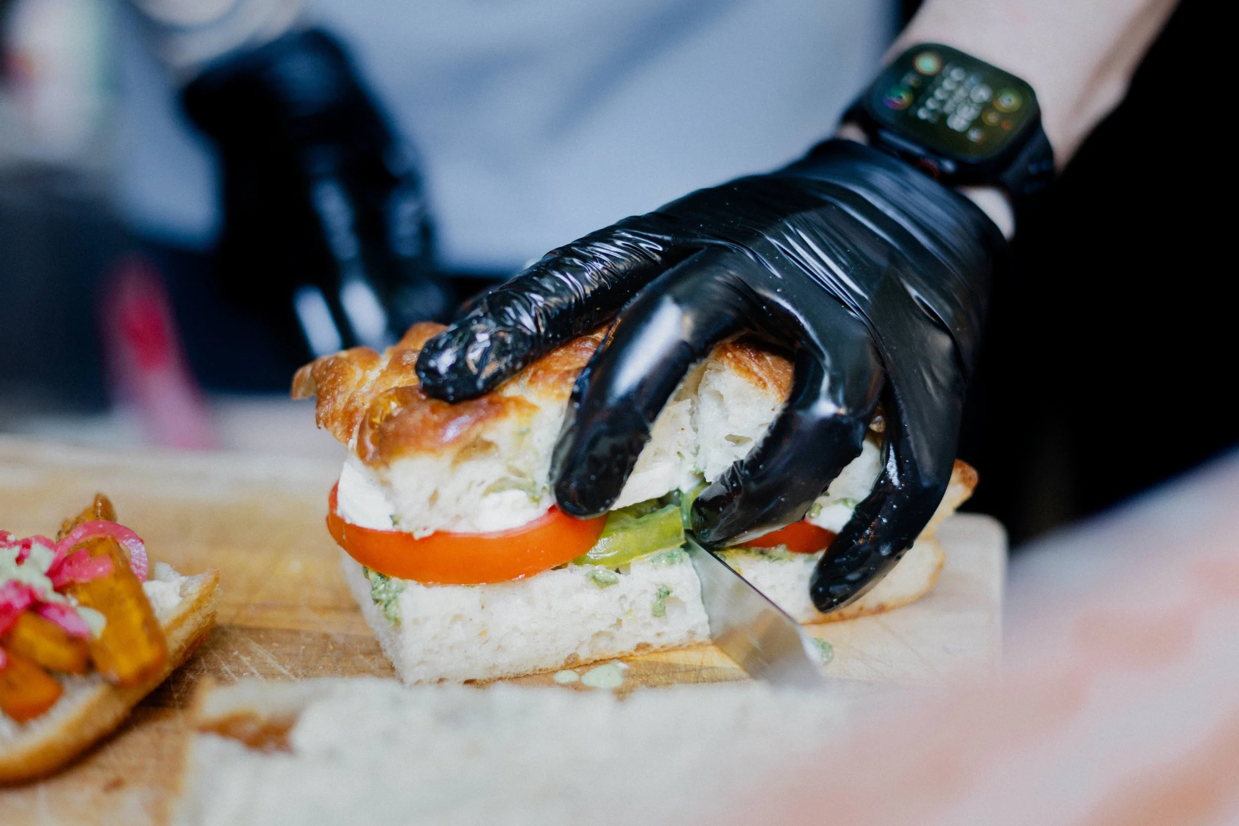 A person's hand wearing a black glove is slicing a sandwich with tomatoes, lettuce, and bread on a wooden cutting board.