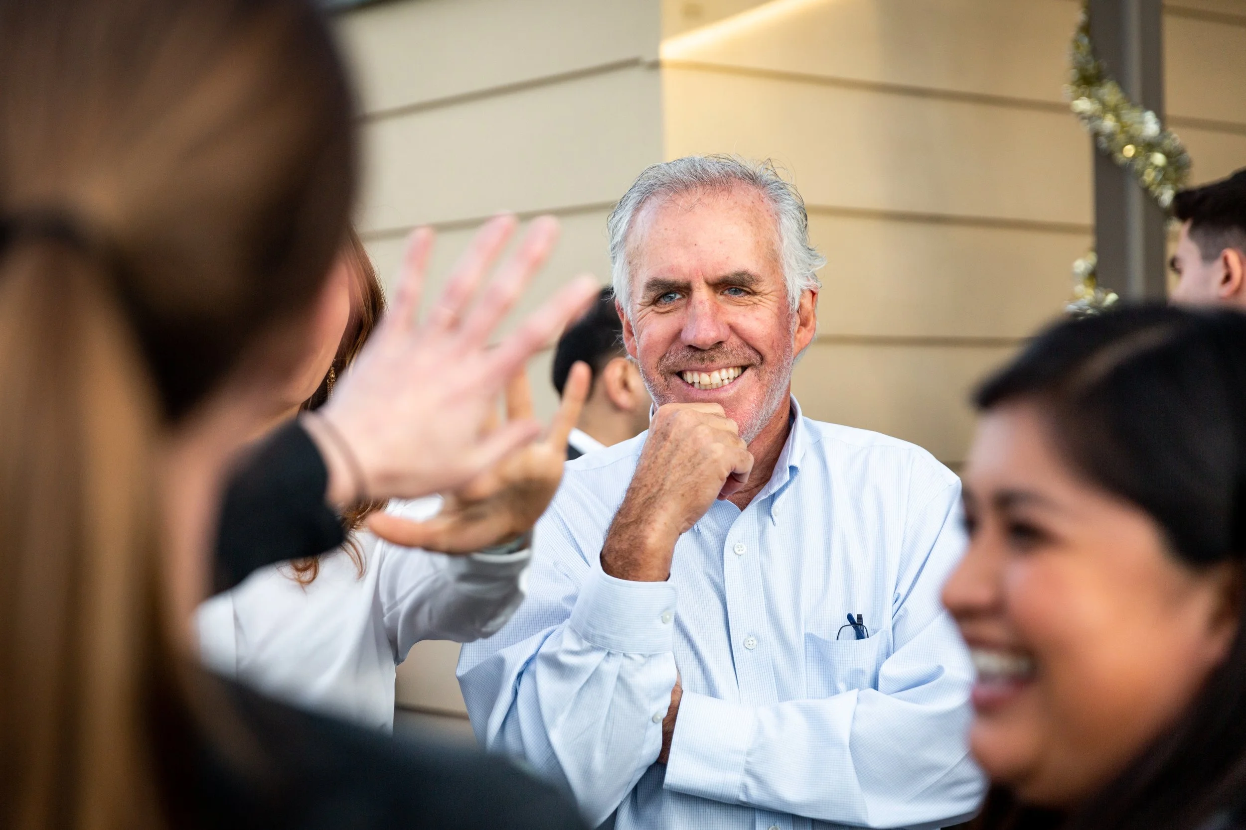 A group of people at a social gathering, with a smiling older man in a light blue shirt in the center, engaging with others in conversation.