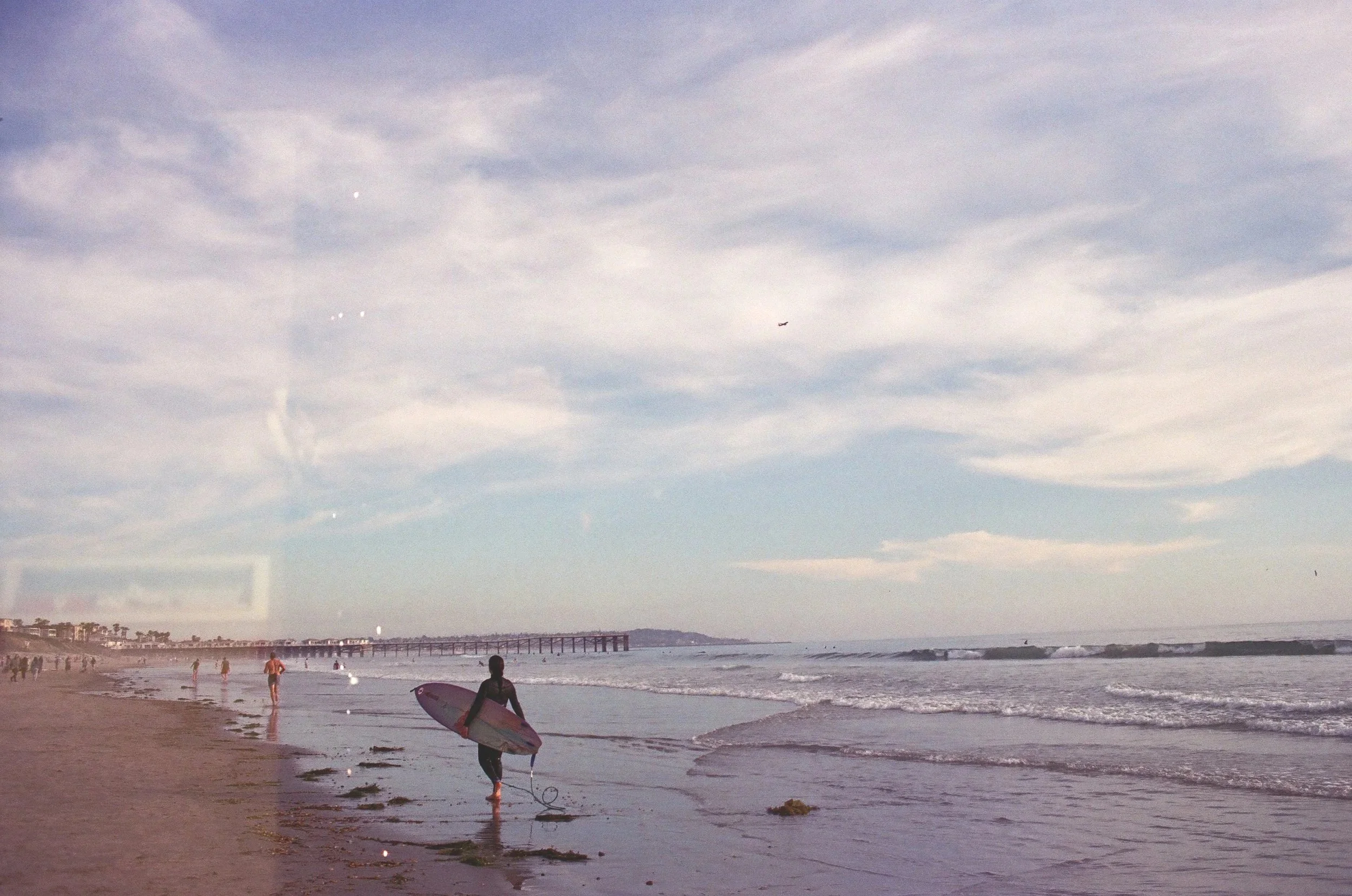A person walking on the beach, holding a surfboard, with a pier extending into the ocean in the background.