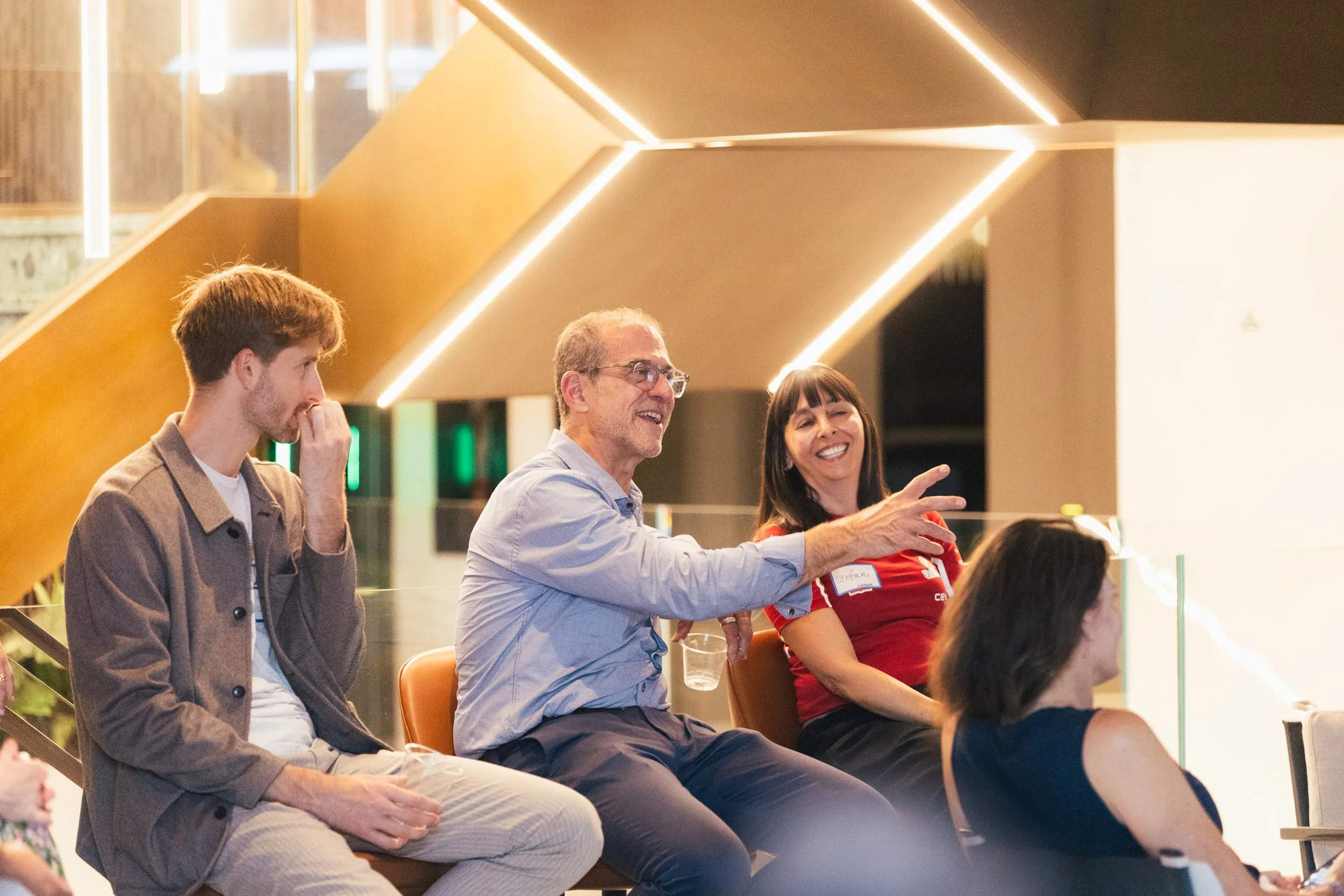 People sitting in a modern indoor space, with three individuals in focus, two of them laughing and engaged in conversation, one person with a thoughtful expression.