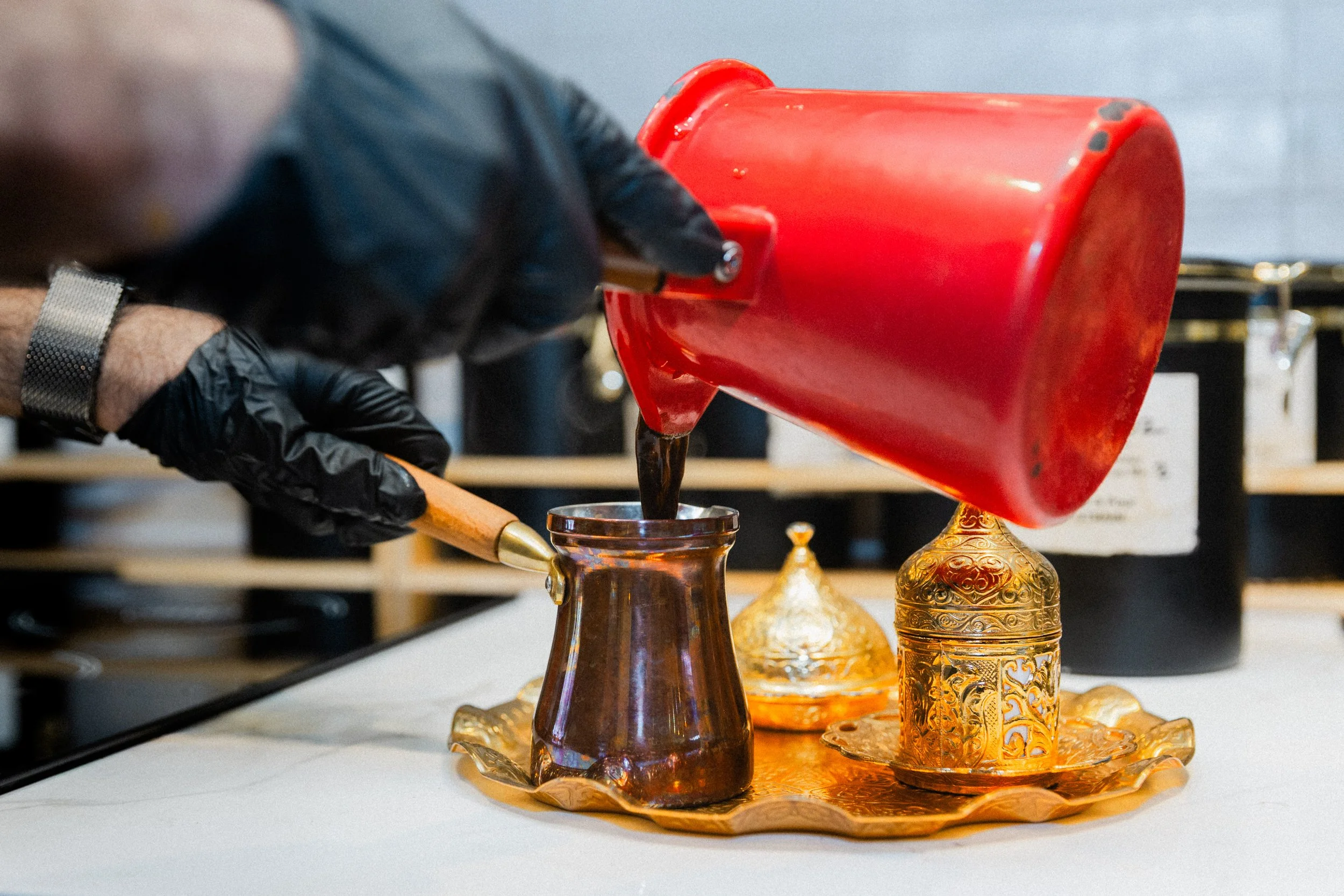 A person wearing black gloves and pouring dark liquid from a red container into a small ornate glass cup.