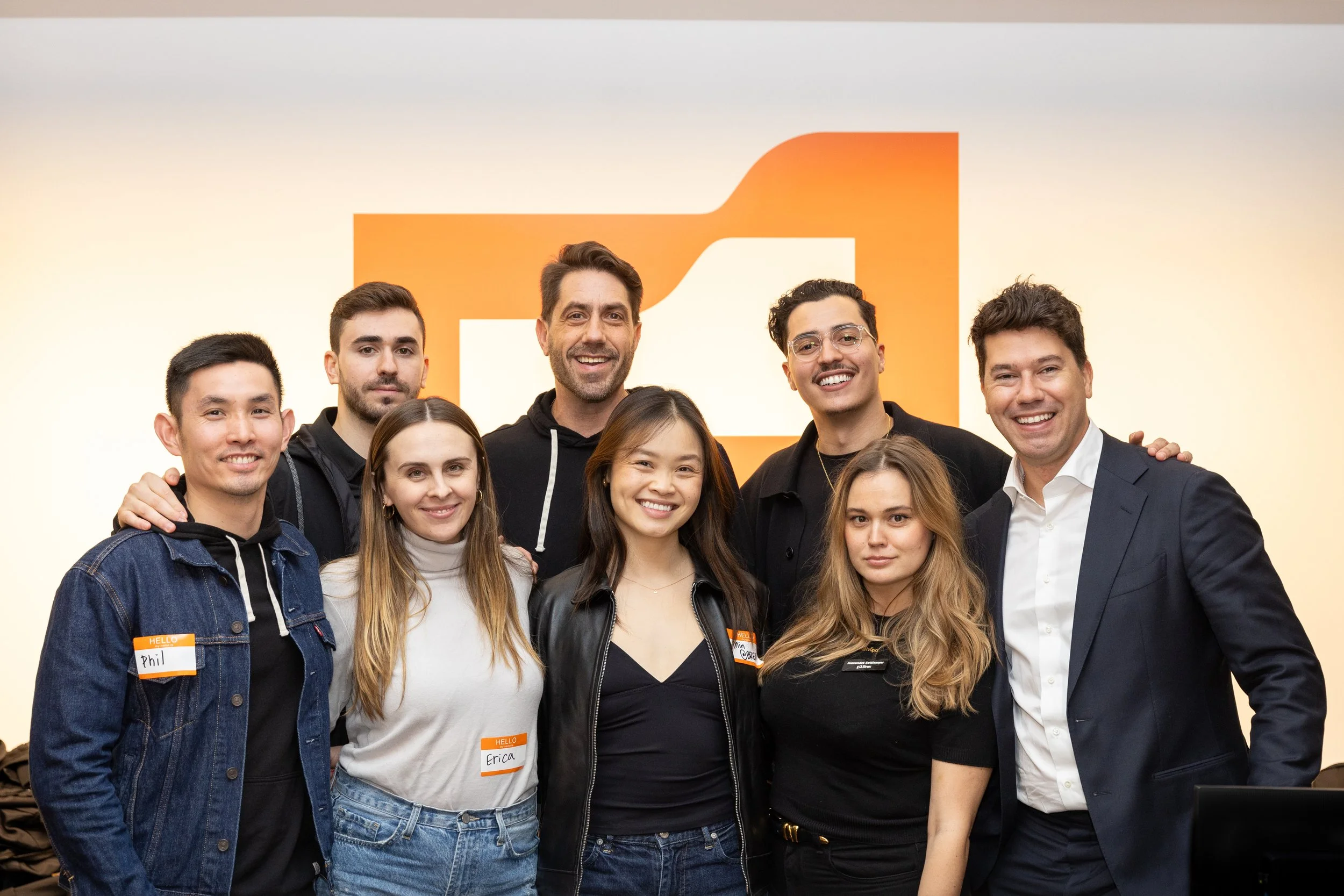 Group of nine diverse young adults smiling and standing together in an indoor setting, with an orange and white geometric logo in the background.