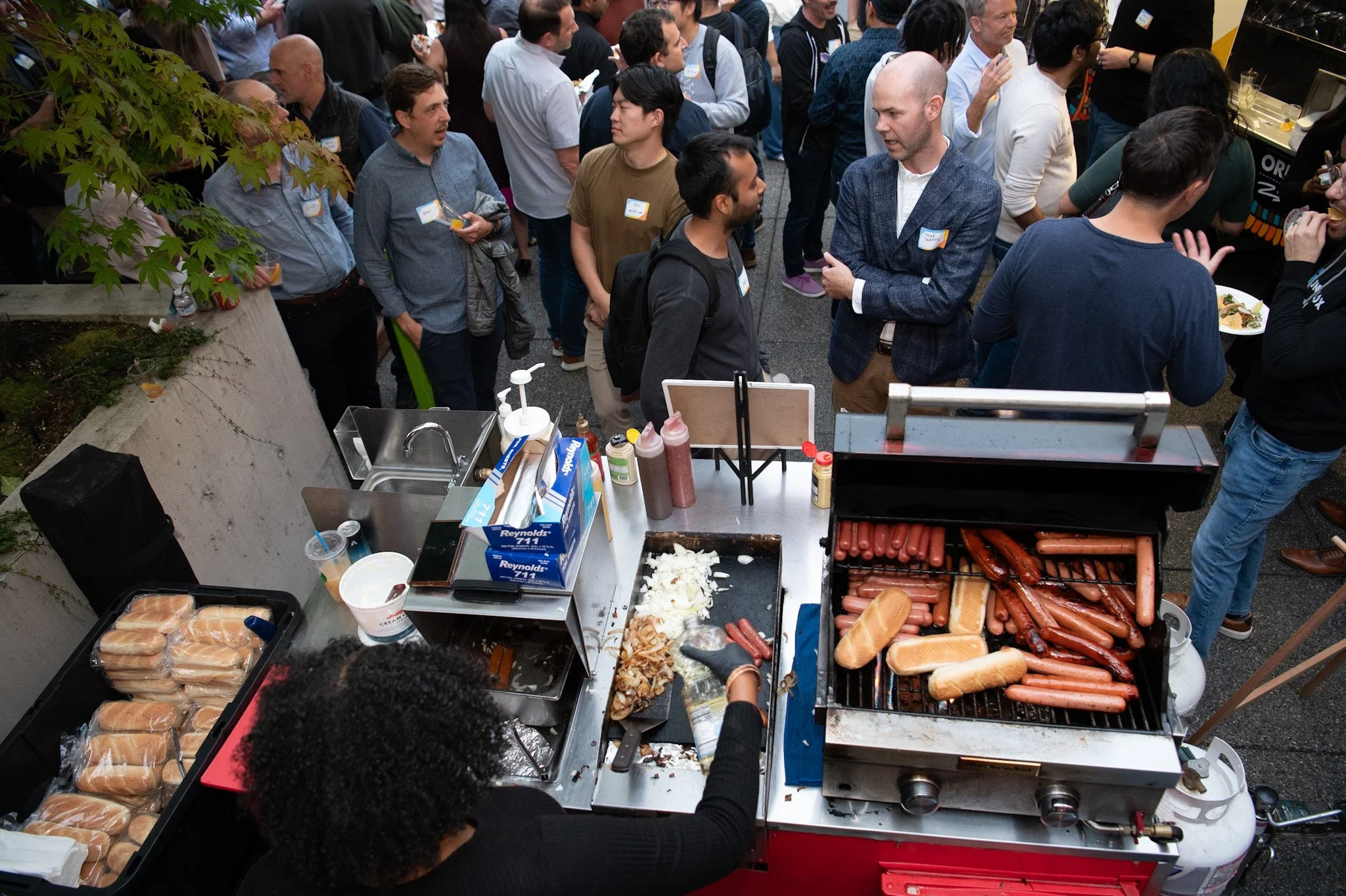 Crowd gathering at an outdoor food stand with hot dogs and buns, people waiting and chatting at a busy event
