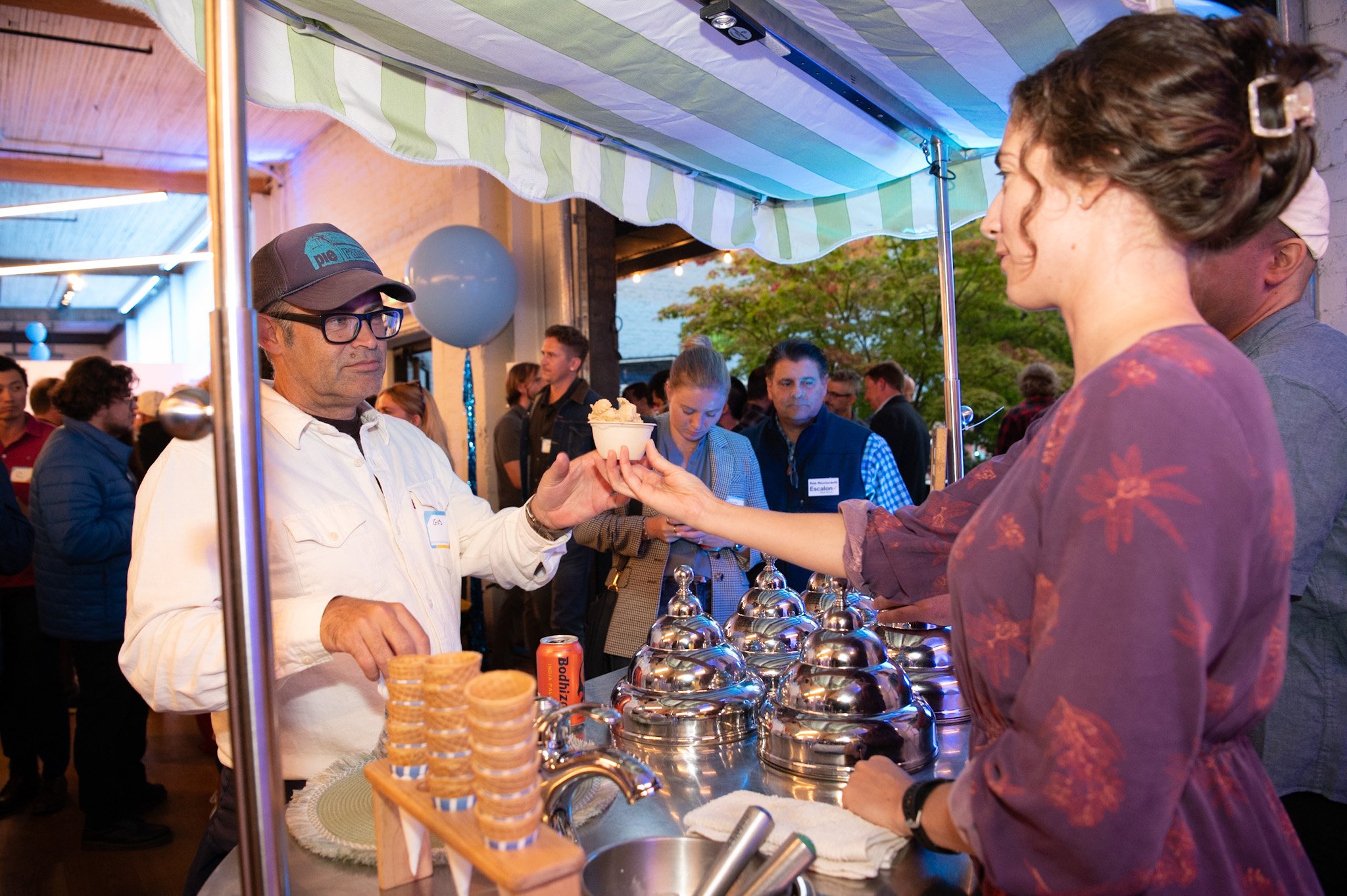A person in a white shirt and cap is receiving an ice cream from a woman at a food stand during an outdoor event.