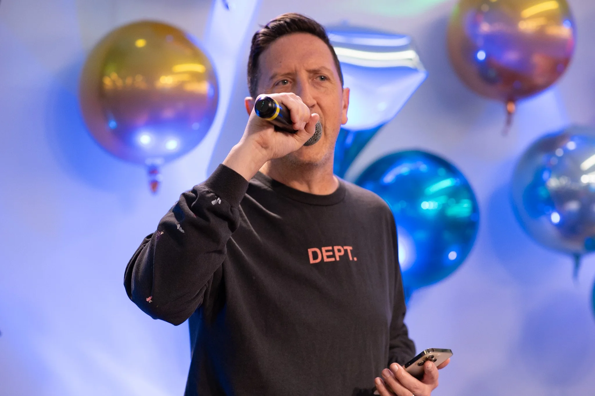 A man singing into a microphone at a party with balloons in the background.