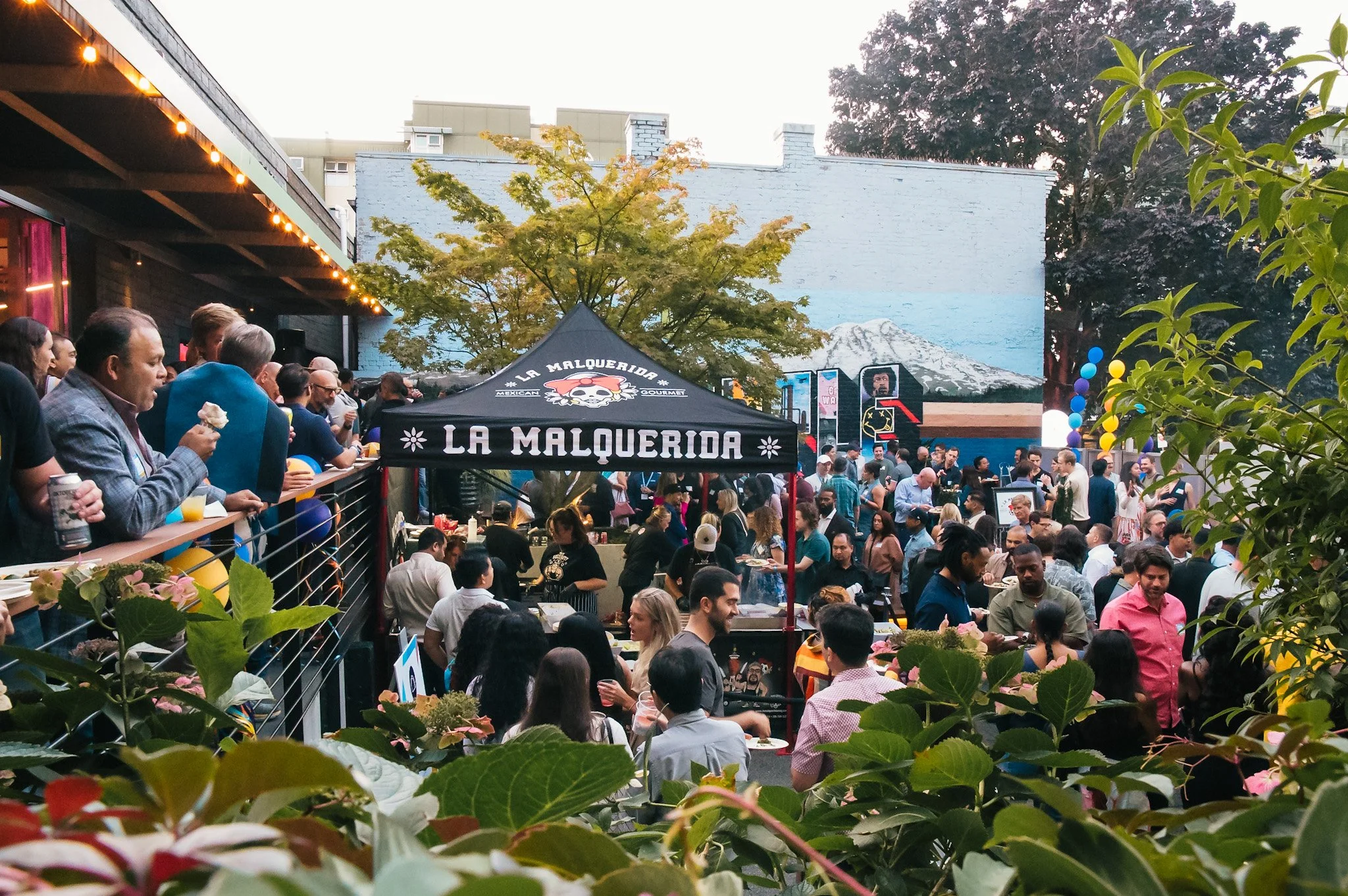Crowd of people at an outdoor event with a black tent labeled 'La Malquerida', some eating, drinking, and socializing. The scene features trees, mural paintings, and string lights.