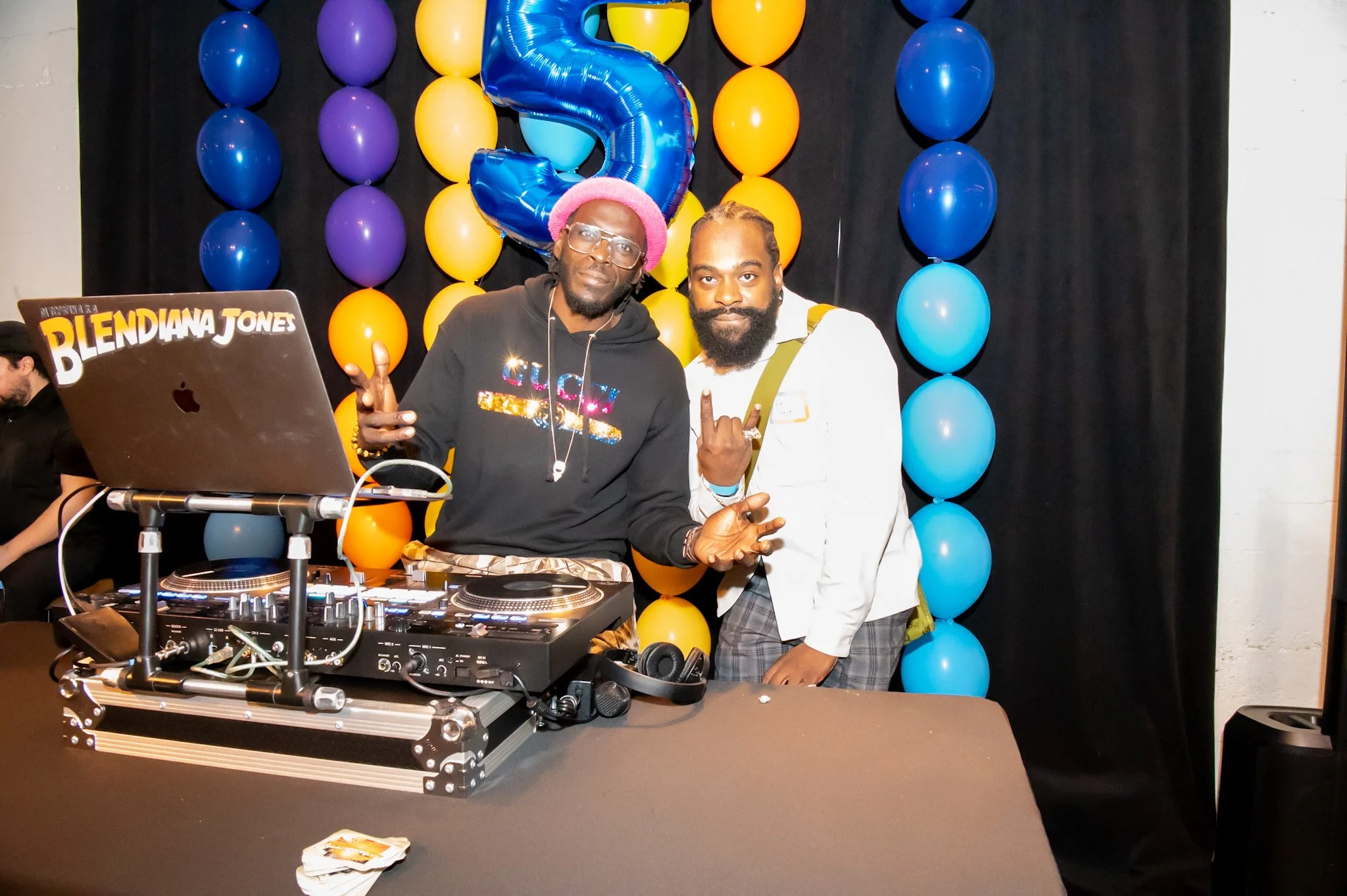 Two men standing behind a DJ setup at a birthday celebration, with blue, purple, orange, and yellow balloons in the background, including a large blue number five balloon.