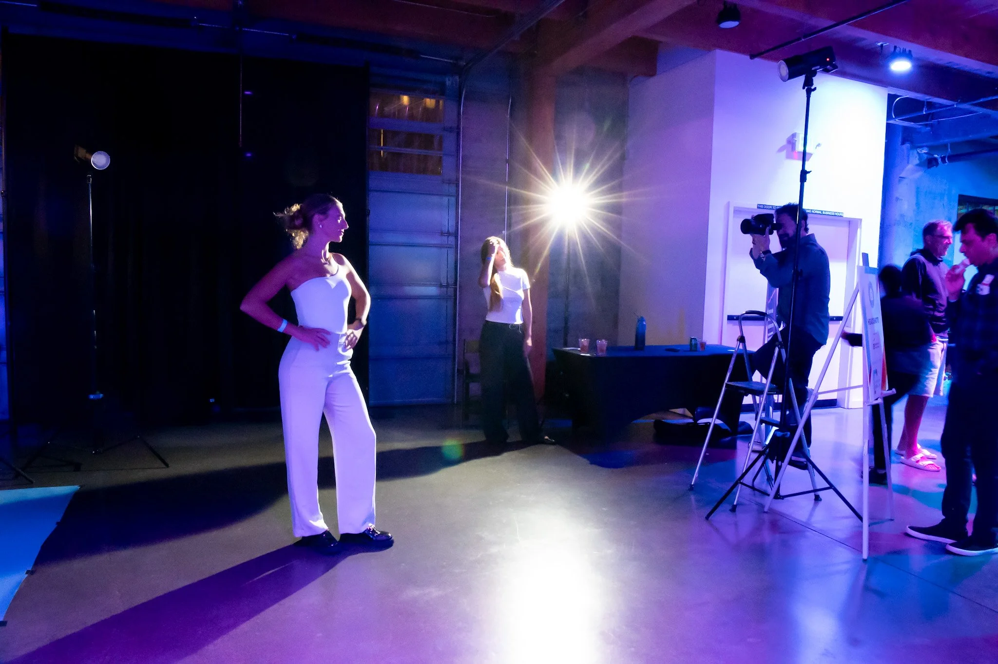 People at a party or event with lighting and photography equipment, with two women in focus, one in a white outfit, and a photographer taking pictures.
