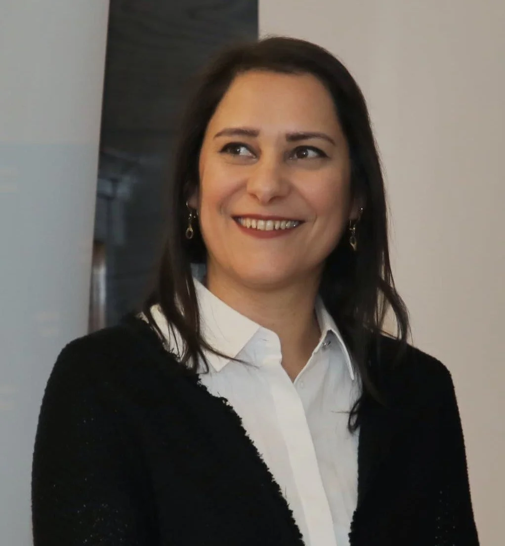 A woman with dark hair smiling, dressed in a white shirt and black cardigan, standing indoors.