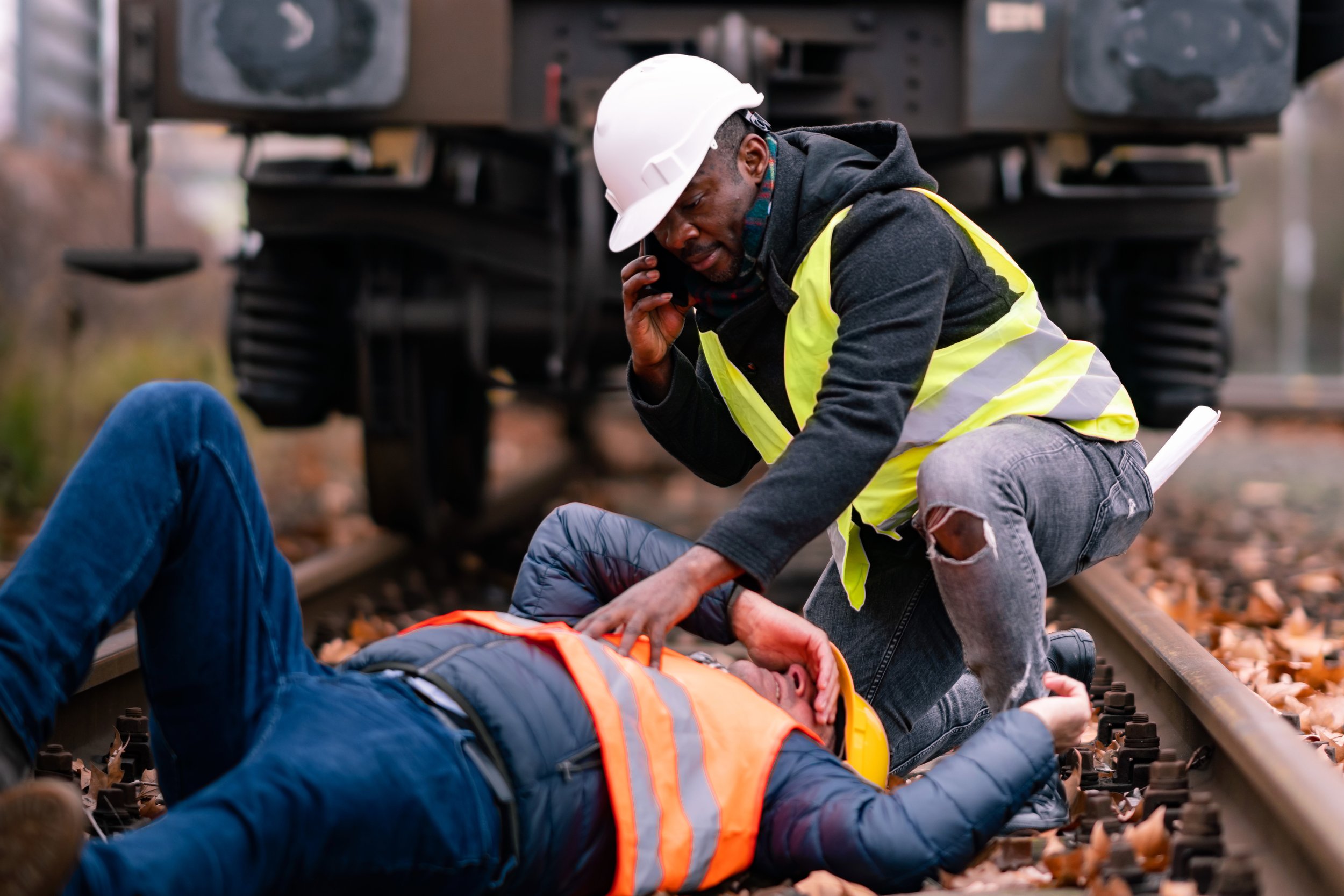 Two workers, one fallen, the other on a phone, near a train on a track