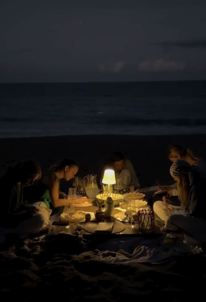 People are having a candlelit dinner on the beach at night, with the ocean in the background.
