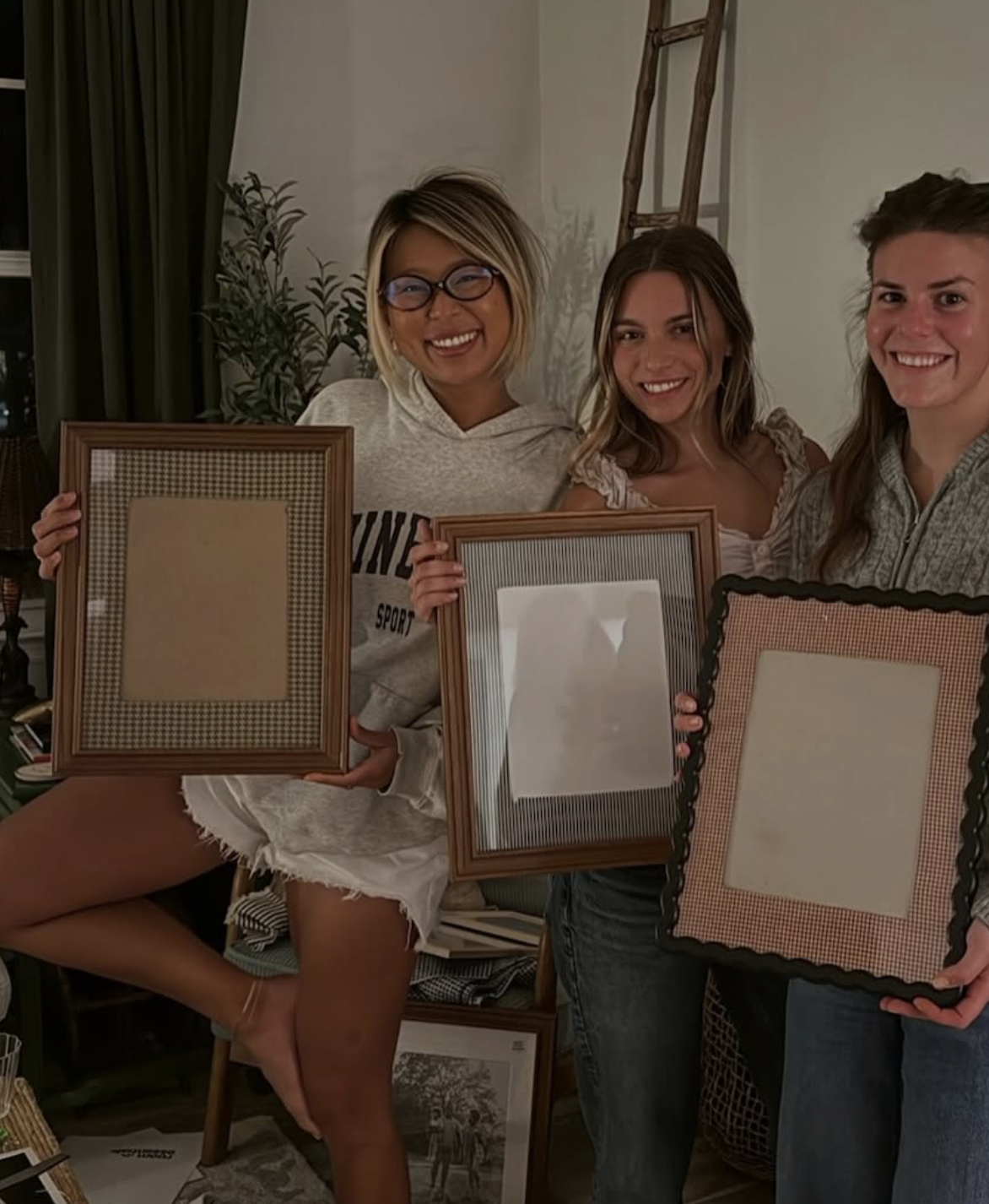 Three women smiling and holding empty picture frames in a cozy indoor setting.