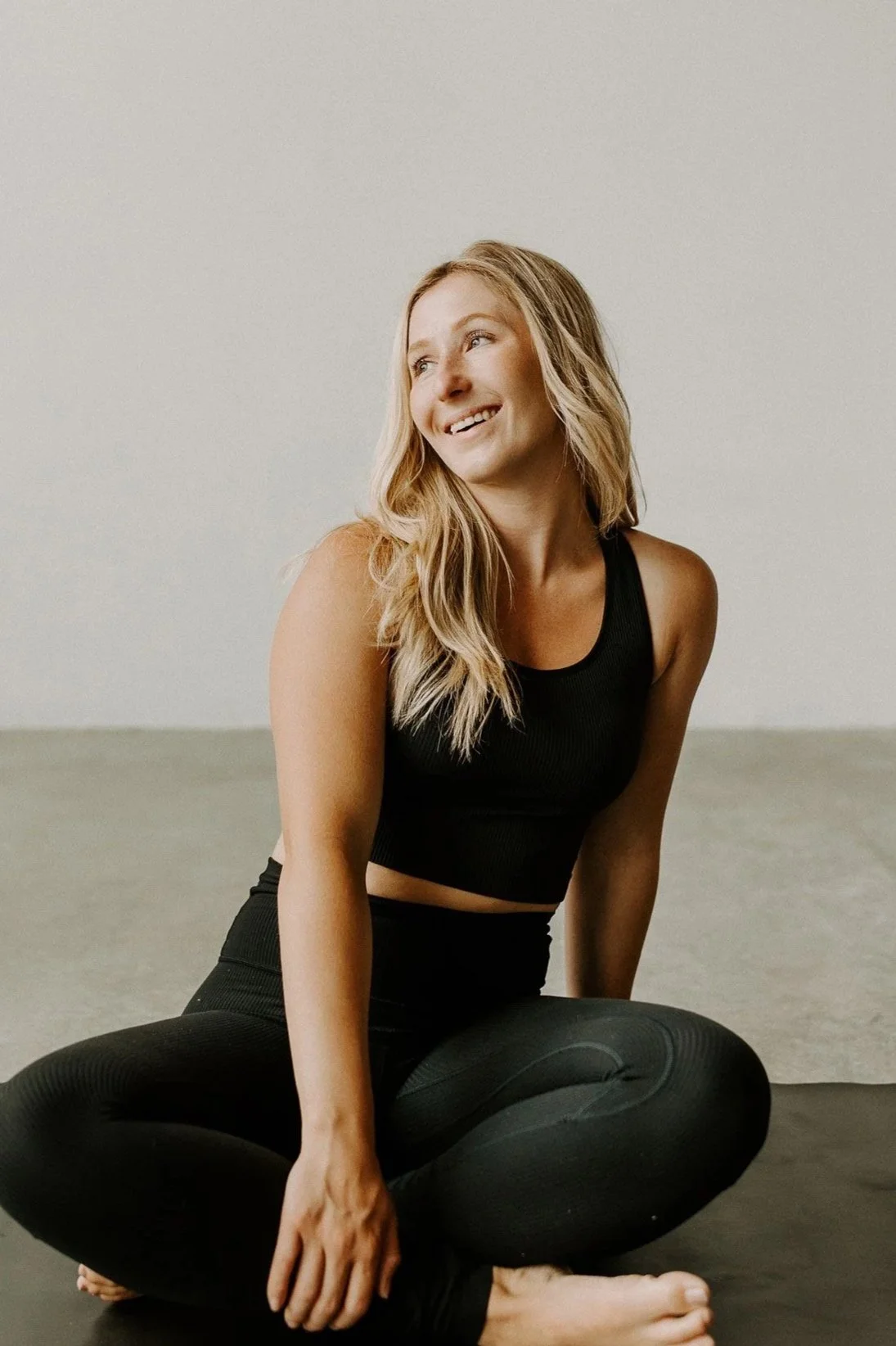 A woman with blonde hair sitting cross-legged on the floor, wearing a black sleeveless workout top and black leggings, smiling and looking to her right against a plain light-colored wall.