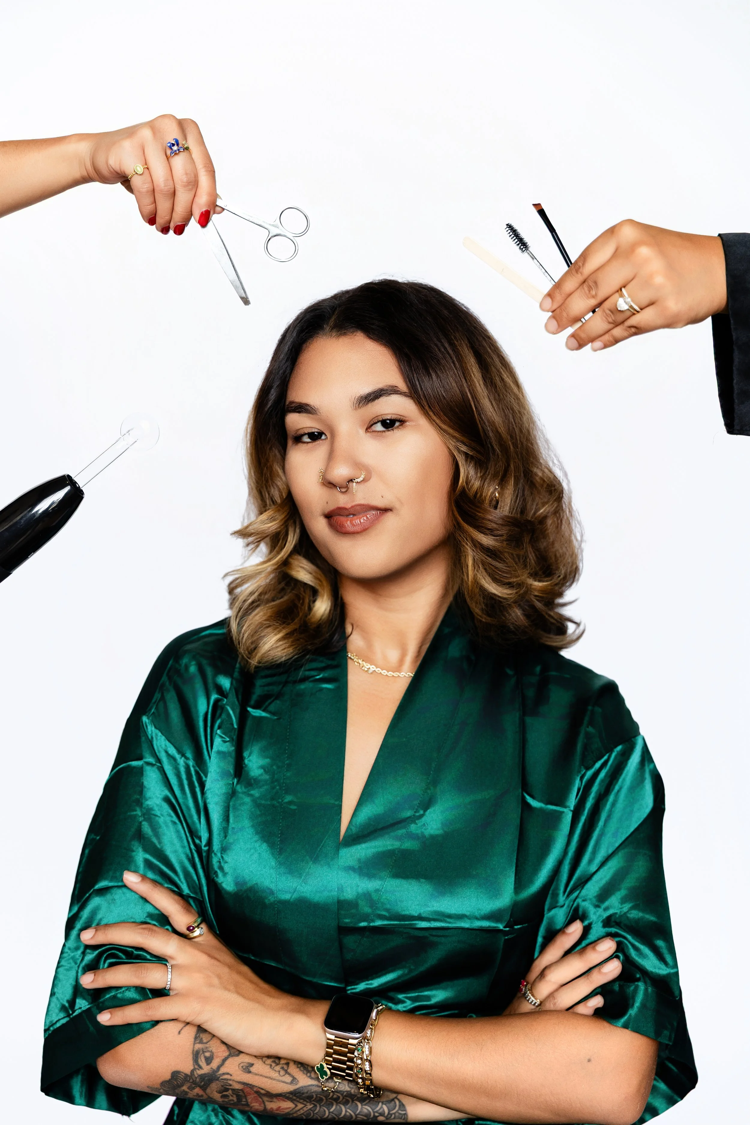 A woman with wavy brown hair and a septum piercing wearing a shiny dark green satin robe, with tattoos on her arms, crossed arms, and various jewelry, surrounded by hands holding hairstyling tools and makeup brushes.