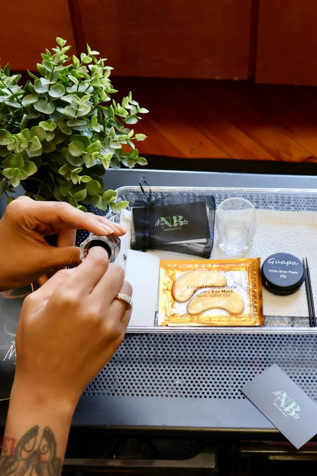 A person's hands are preparing for a beauty treatment on a metal tray, with grooming items, including a face mask, eye mask, and brow paste, with a green potted plant and a wooden wall in the background.