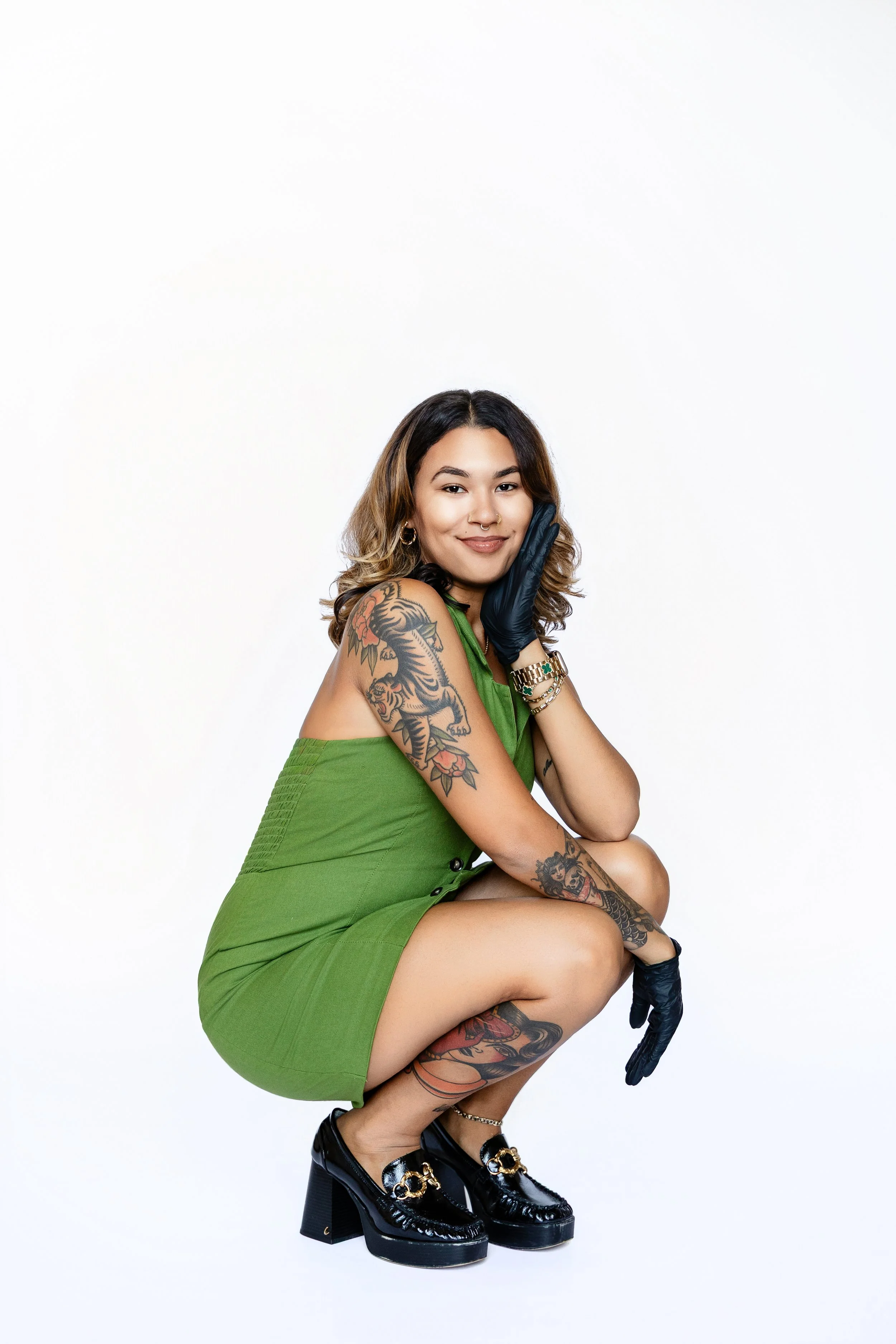 A woman with tattoos wearing a green dress and black chunky heels, squatting and smiling while holding her face with one hand and resting her elbow on her knee, posing against a plain white background.