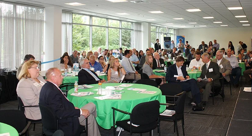 Business conference with attendees seated at round tables in a room with large windows and natural light.