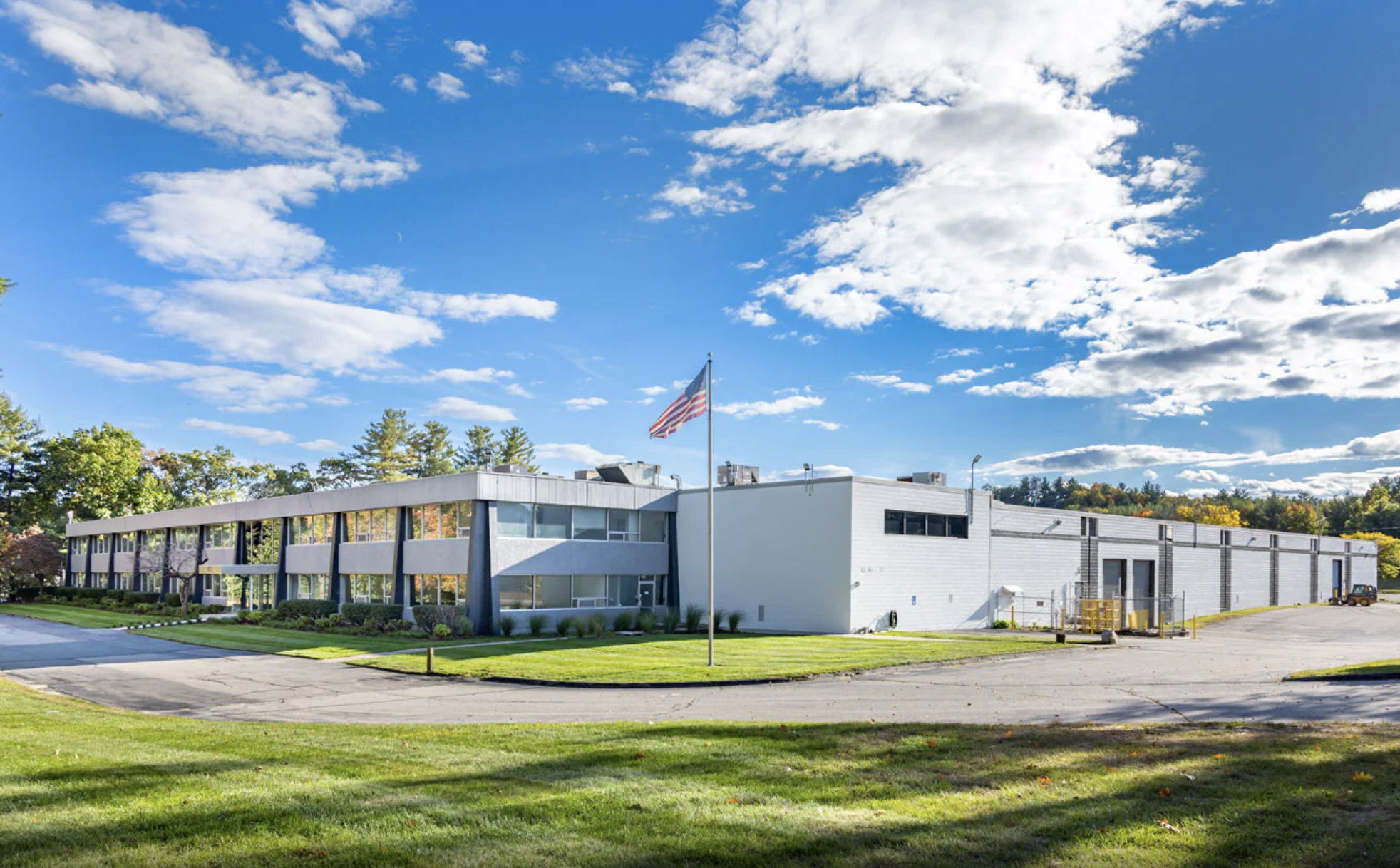 Modern building with white and gray exterior walls, large windows, surrounded by green grass and trees, under a partly cloudy blue sky with scattered clouds and an American flag on a flagpole.