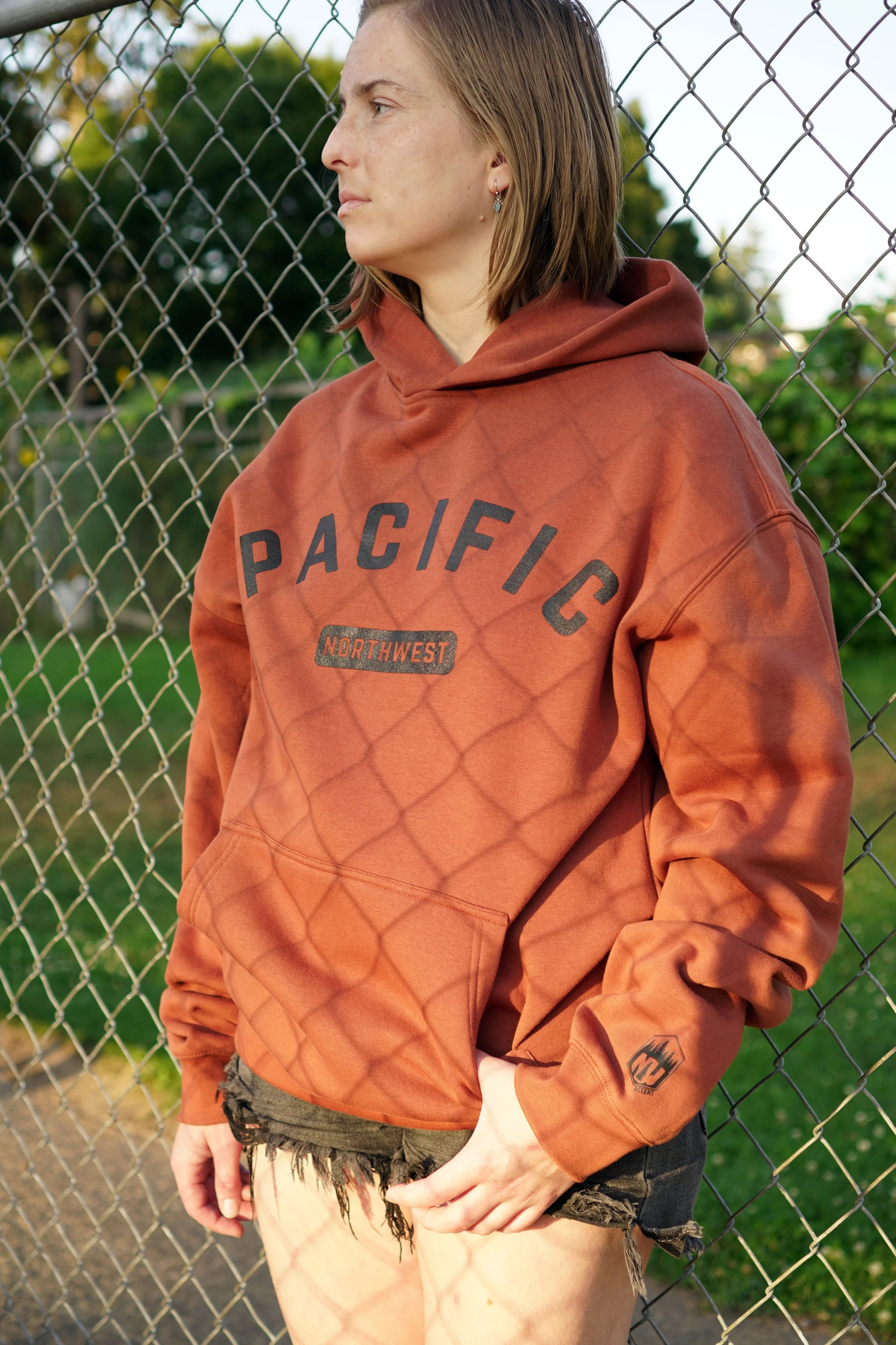 A woman leaning against a chain-link fence outdoors, wearing an oversized rust-colored hoodie with the words 'PACIFIC' and 'NORTHWEST' printed on the front, and frayed denim shorts.