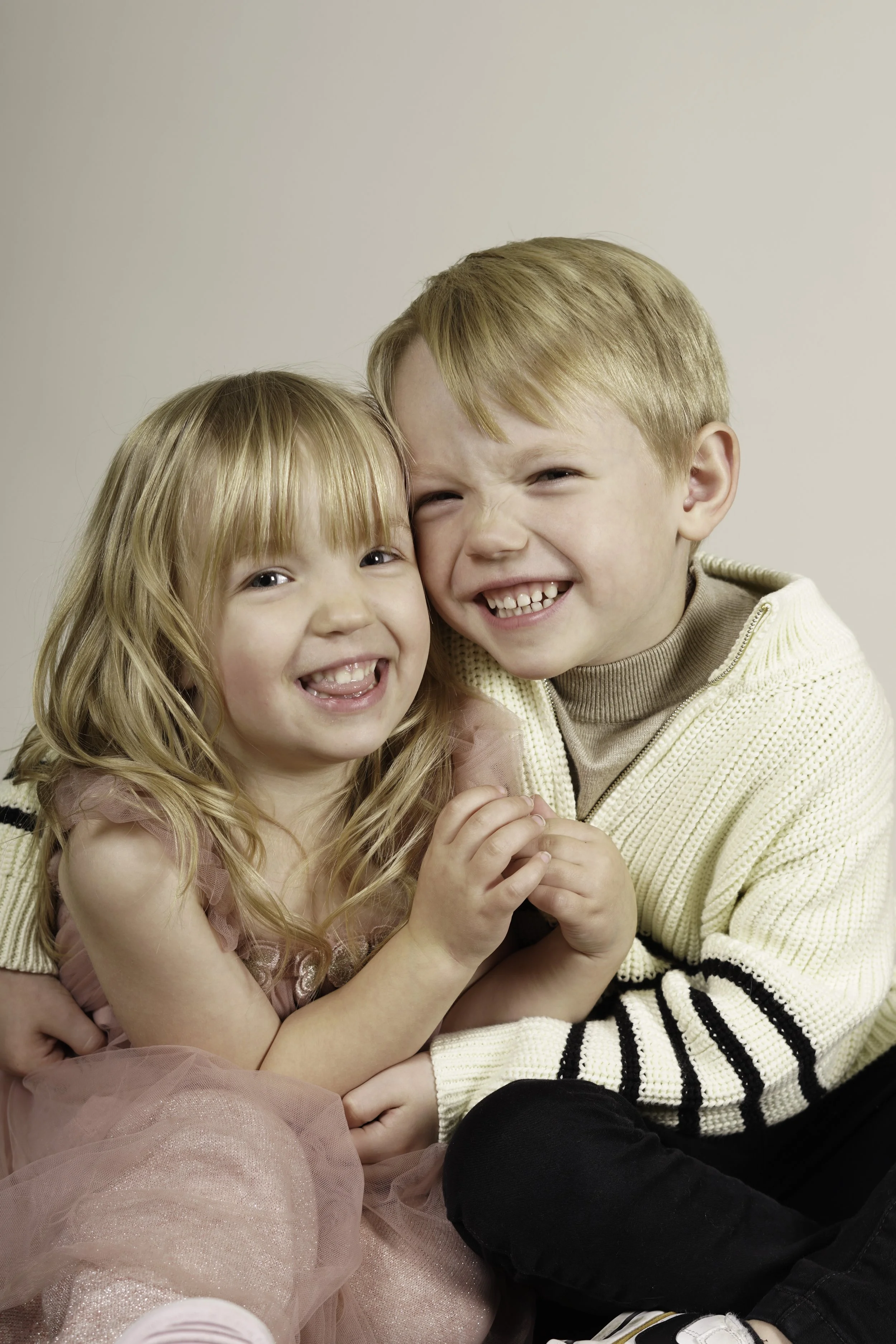Two young children, a girl and a boy, are smiling and hugging each other. The girl has long blonde hair and is wearing a pink dress, while the boy has short blonde hair and is dressed in a cream sweater with black and white stripes.