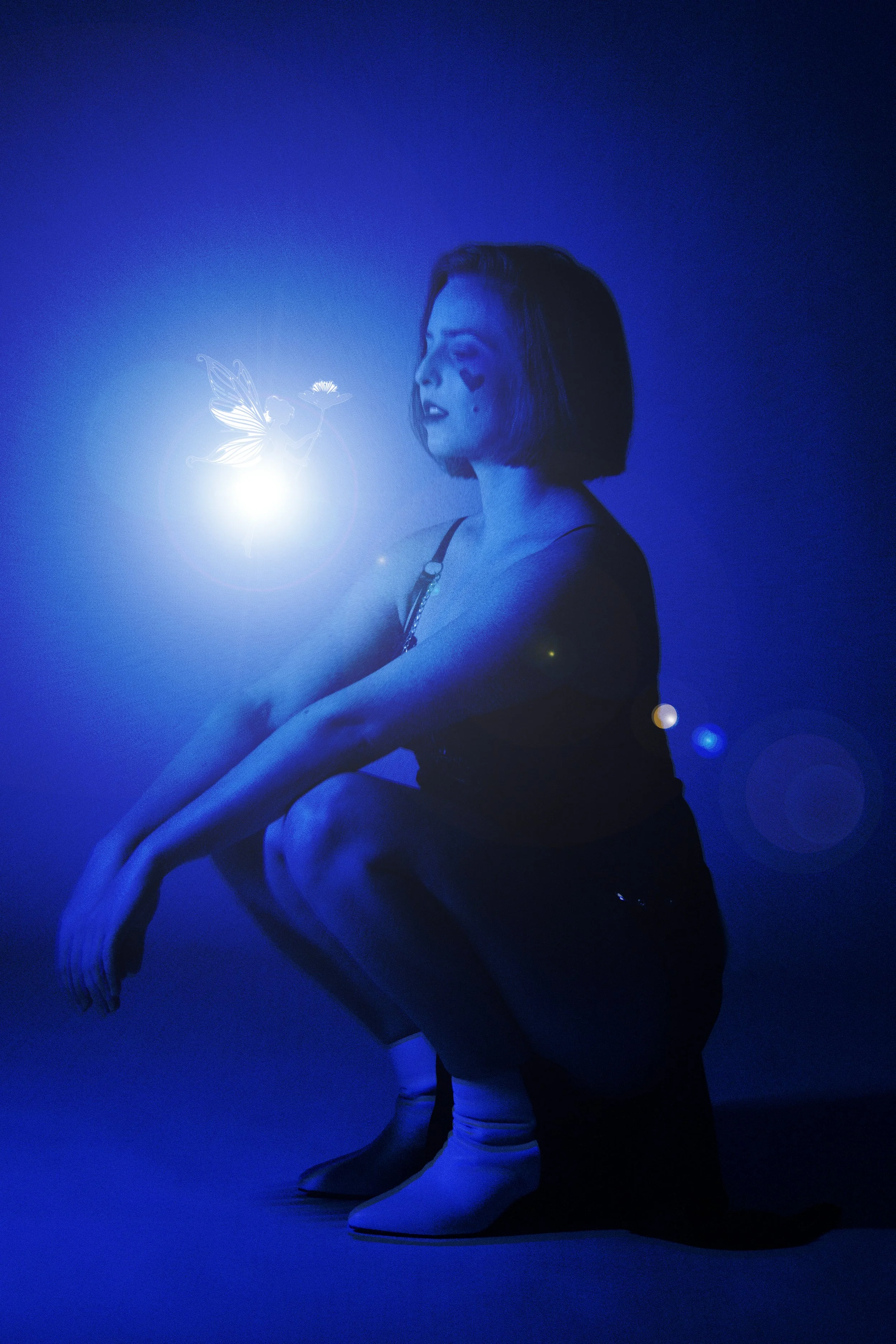 A woman with short hair and a dark tank top squats in a blue-lit room, with a glowing fairy and a flower near her face.