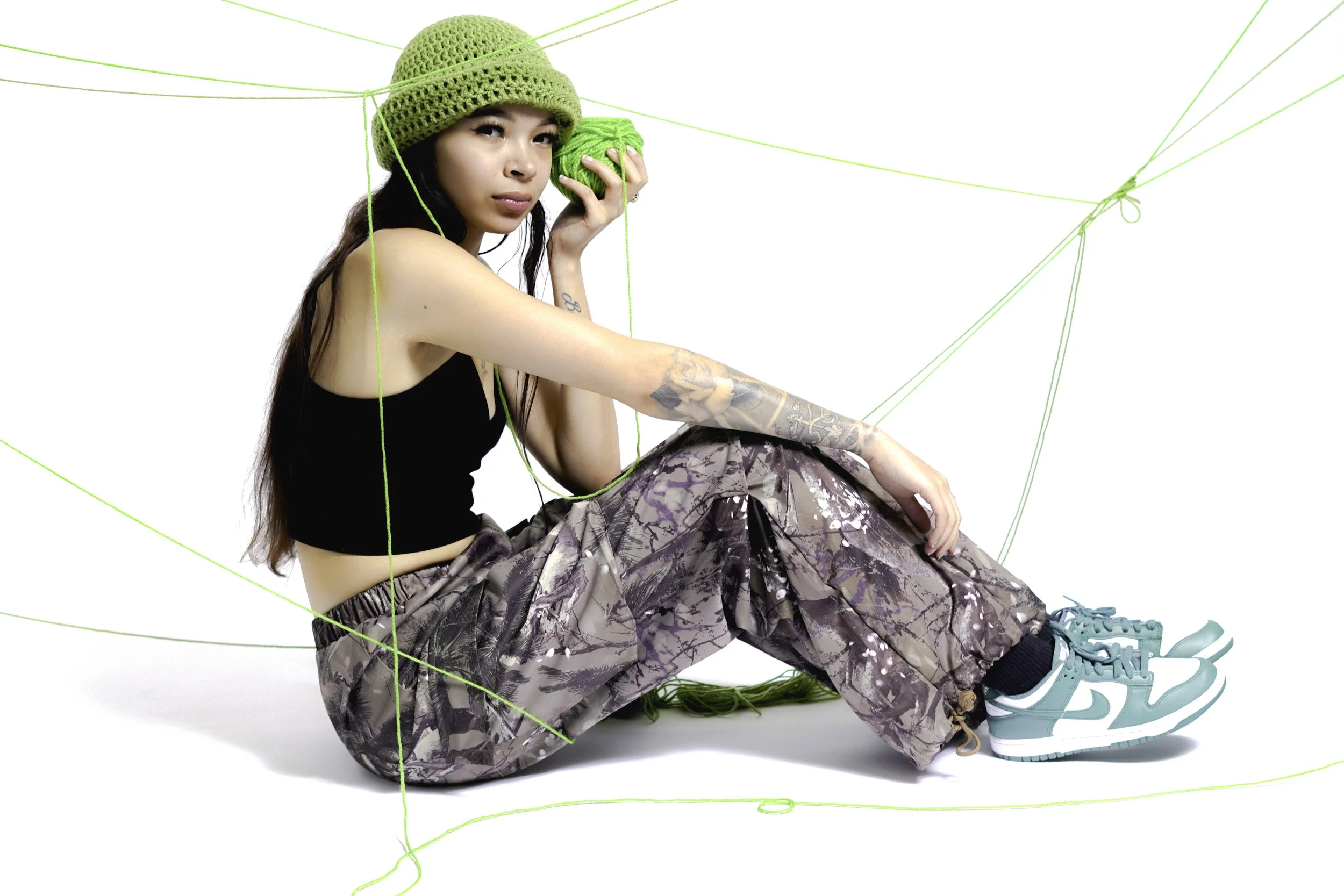 Young woman sitting on the floor, holding a ball of green yarn with knitting needles attached, wearing a green knit hat, black crop top, camouflage baggy pants, and teal sneakers, with green yarn strings around her.