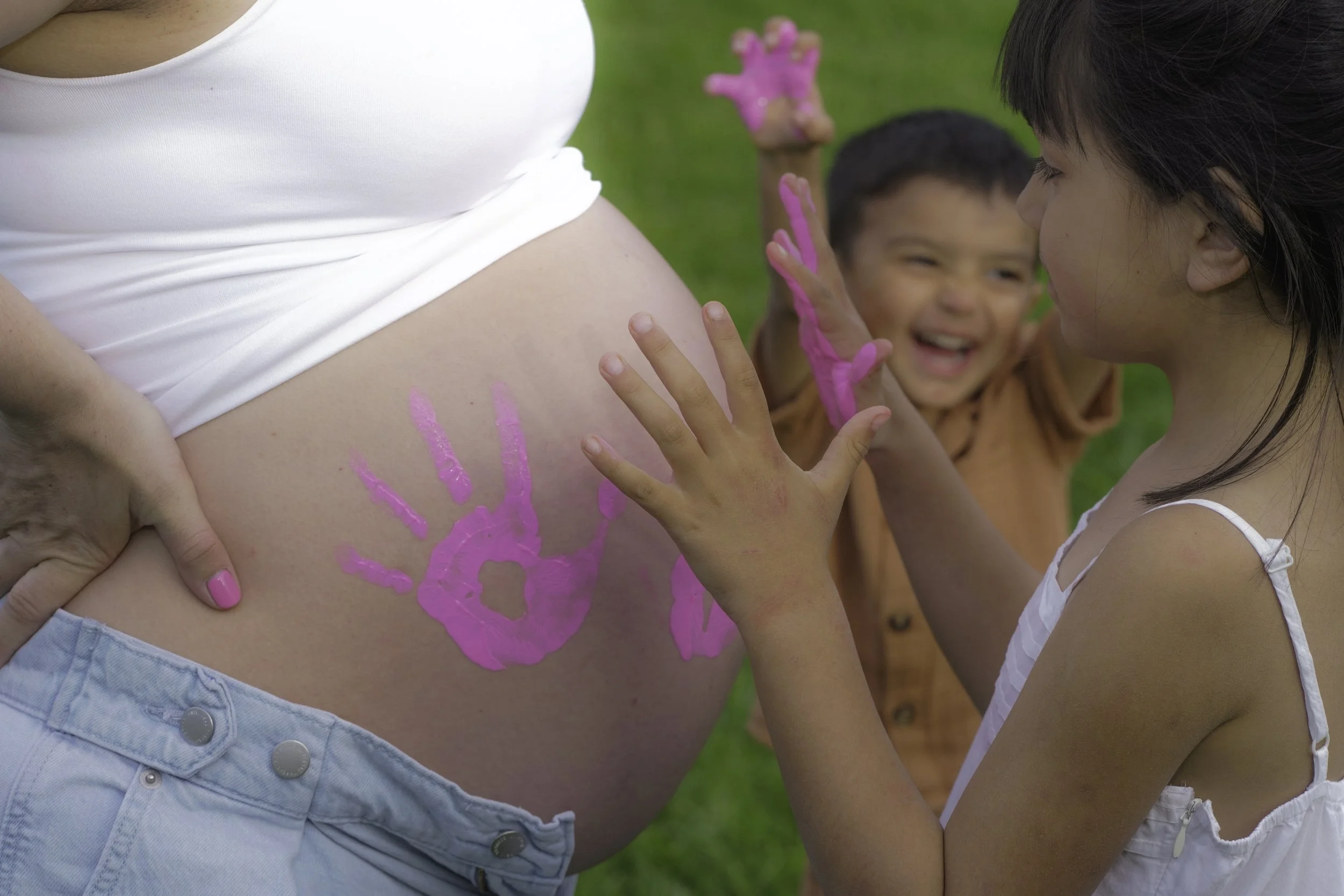 Children painting handprints on a pregnant woman's belly outside in a park.