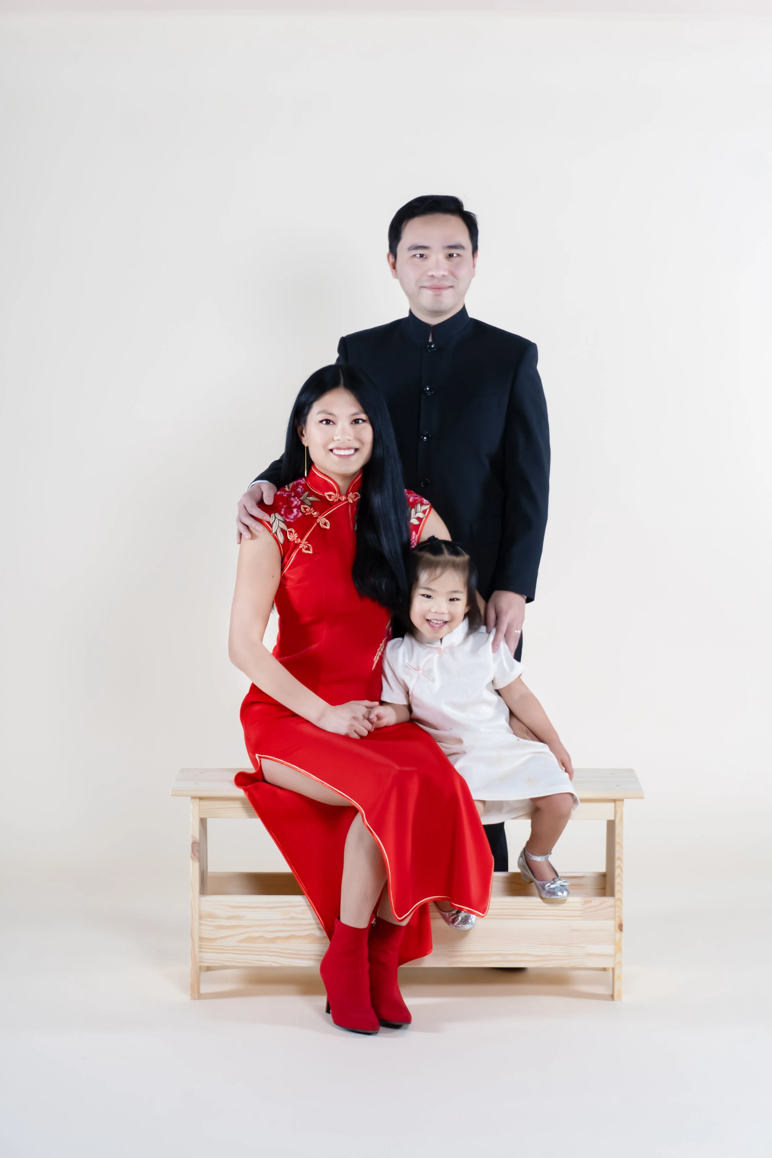 A family portrait featuring a woman in a red traditional dress, a man in a black suit, and a young girl in a white dress, with the woman and girl seated on a wooden bench and the man standing behind them against a plain white background.
